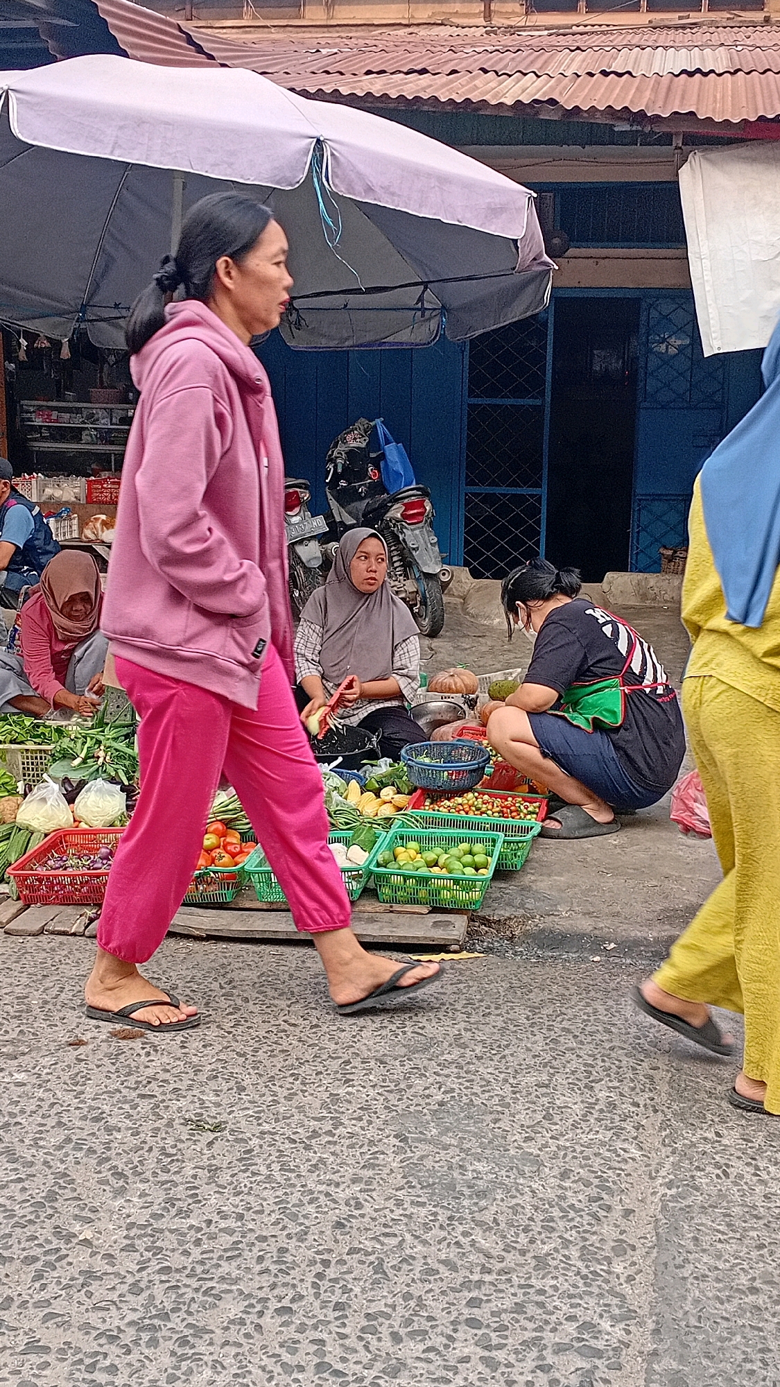 A street market scene in Jambi, Indonesia, features several people interacting with produce. In the foreground, a woman in a pink hooded jacket and bright pink pants walks from left to right, wearing flip-flops. To her right, a woman wearing a grey hijab and patterned shirt sits behind a display of vegetables and fruits, holding a knife and a piece of produce. She appears to be engaged in preparing food. Further to the right, another woman wearing a black t-shirt with a graphic design and a green apron is crouched down, looking at items on the ground. She is also wearing a head covering and dark shorts. In the background, a motorcycle is parked. The market appears to be set up along a street or pathway, with corrugated metal roofing and blue storefronts visible in the background. The ground is paved with rough concrete.