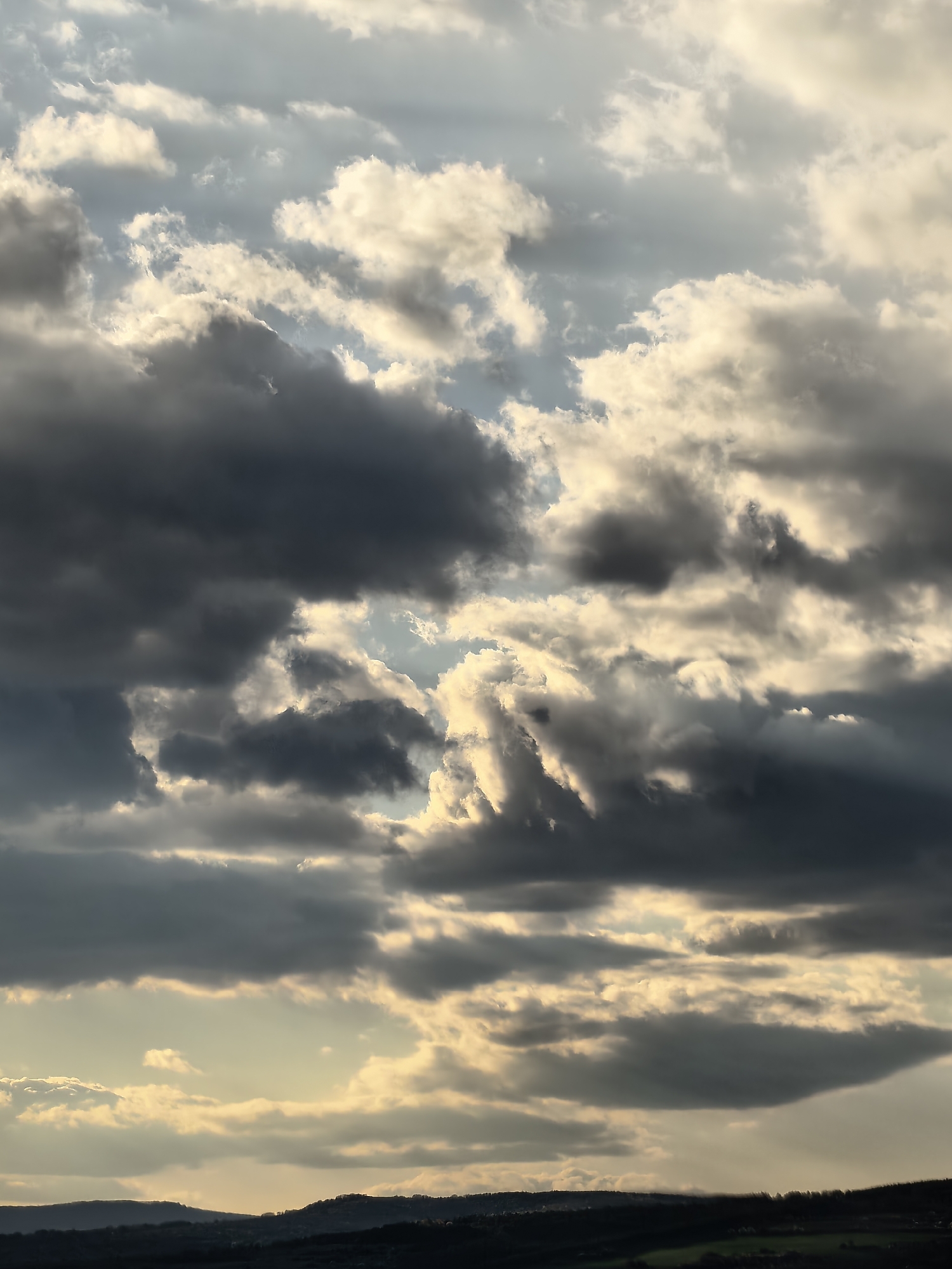 The image shows a cloudy sky above a silhouetted landscape. The sky is filled with a mix of dark, grey storm clouds and lighter, sunlit clouds, with patches of pale blue visible in between. The sun is shining through some of the clouds, creating a golden glow. The landscape at the bottom of the image is dark and indistinct, with rolling hills and trees silhouetted against the sky. The location is in the city of Unknown, Unknown.