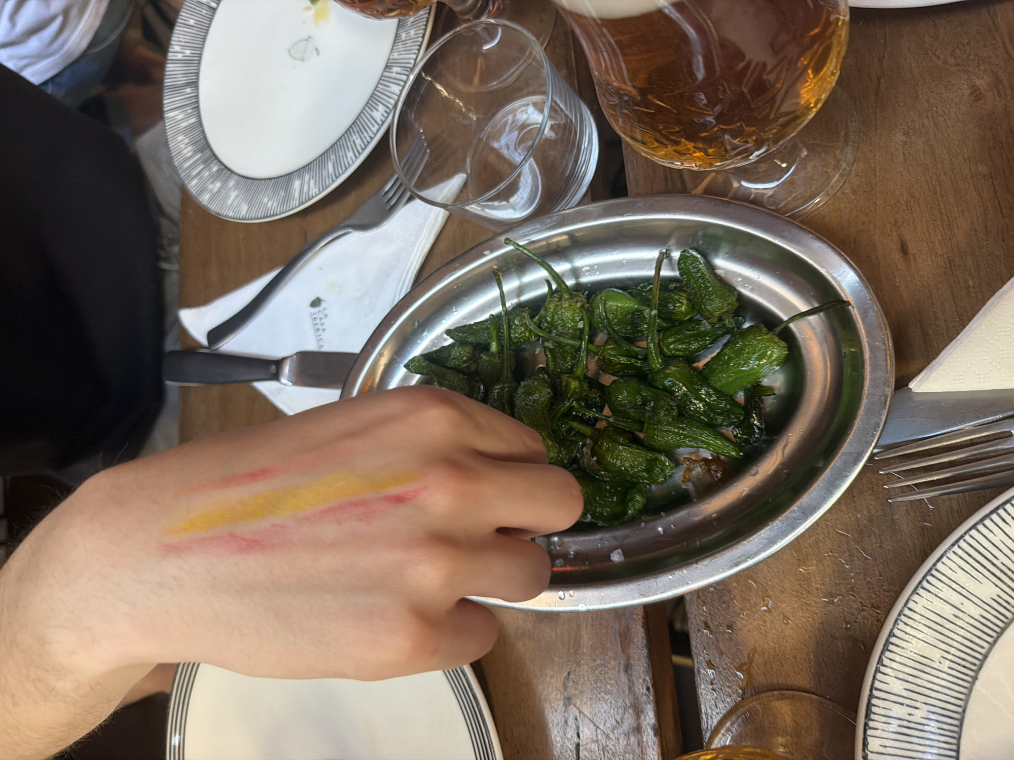 A person's hand with red and yellow streaks of paint on the back of it is reaching into a metal bowl filled with green peppers, likely Padrón peppers, sprinkled with salt. The bowl is placed on a wooden table, alongside white plates with black and white striped rims, cutlery, and a glass with a beverage. The scene appears to be a casual dining setting, possibly a restaurant in Milan, Italy, suggested by the presence of tableware and drinks.