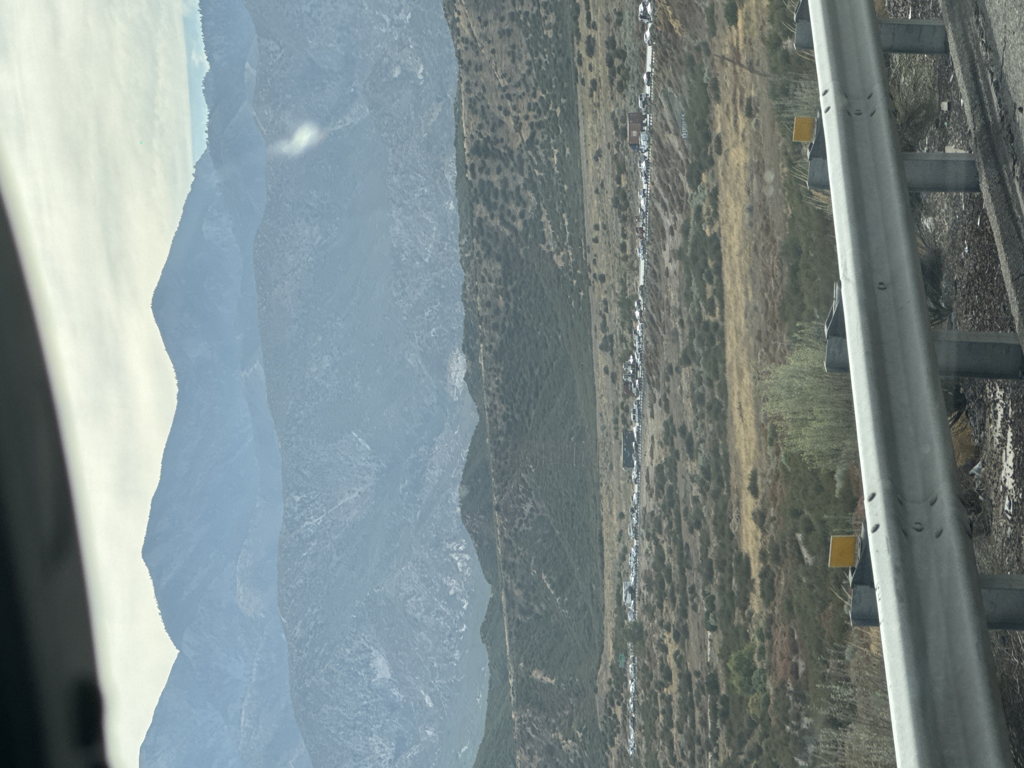 An aerial view captures a traffic jam on a highway in Oak Hills, California. The highway is lined with numerous cars and trucks, indicating a significant traffic backup. The surrounding landscape is characterized by rolling hills covered in dry brush and sparse vegetation, typical of the arid environment of Southern California. In the background, several layers of mountains, possibly the San Bernardino Mountains, are visible, partially obscured by atmospheric haze. The sky is overcast with a few patches of light. The foreground shows a section of a highway guardrail, suggesting the photo was taken from a vehicle moving alongside the traffic.