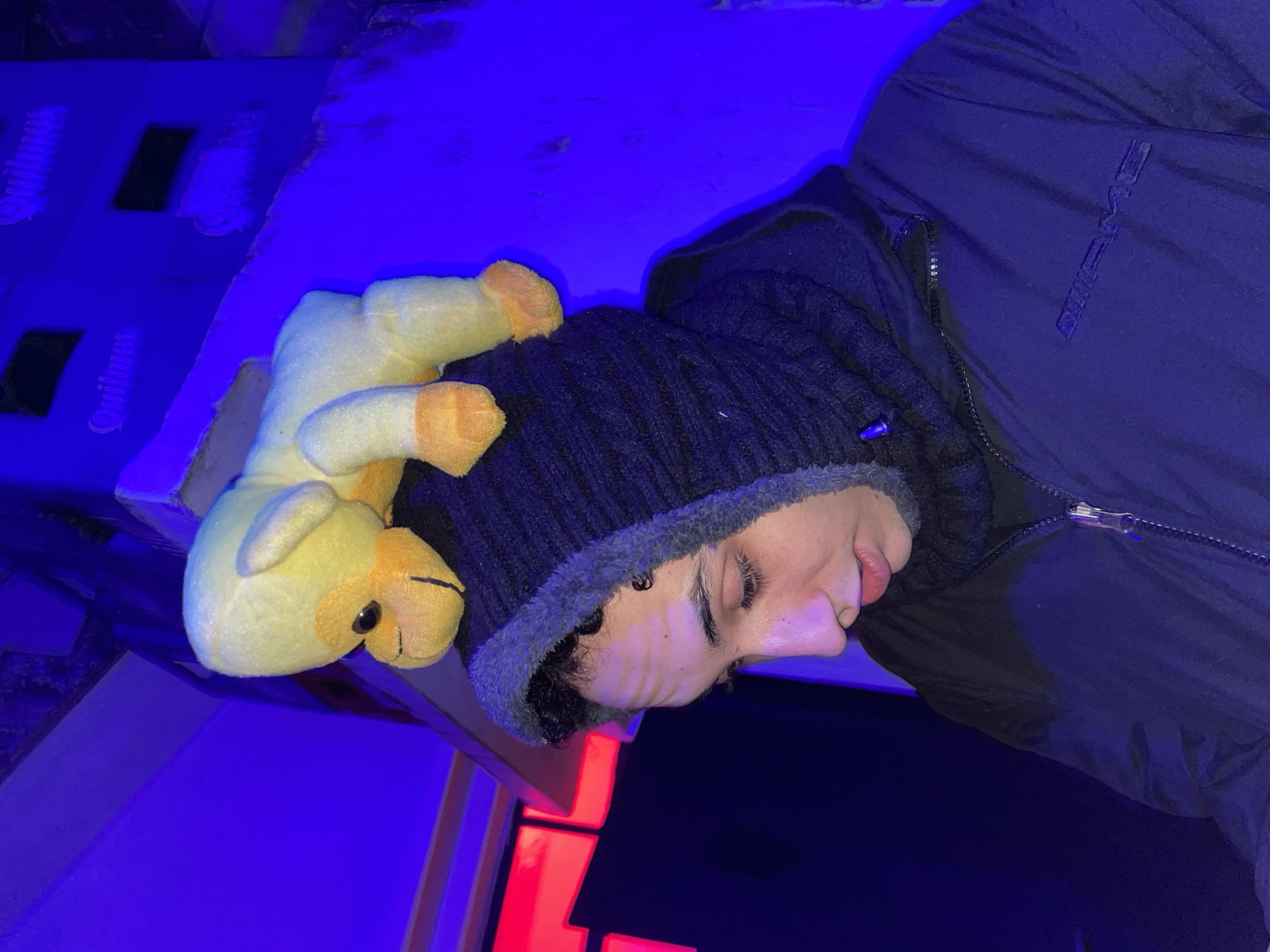 A sideways indoor shot shows a young man wearing a knit cap and a dark jacket. He is lying down with a stuffed, yellow camel resting on his head. Beside him are two blue Quilmes beer cases stacked on top of each other. Part of a red neon sign is visible. The scene appears to be in Argentina due to the Quilmes beer branding. There is no clear indication within the image that the location is Buenos Aires.