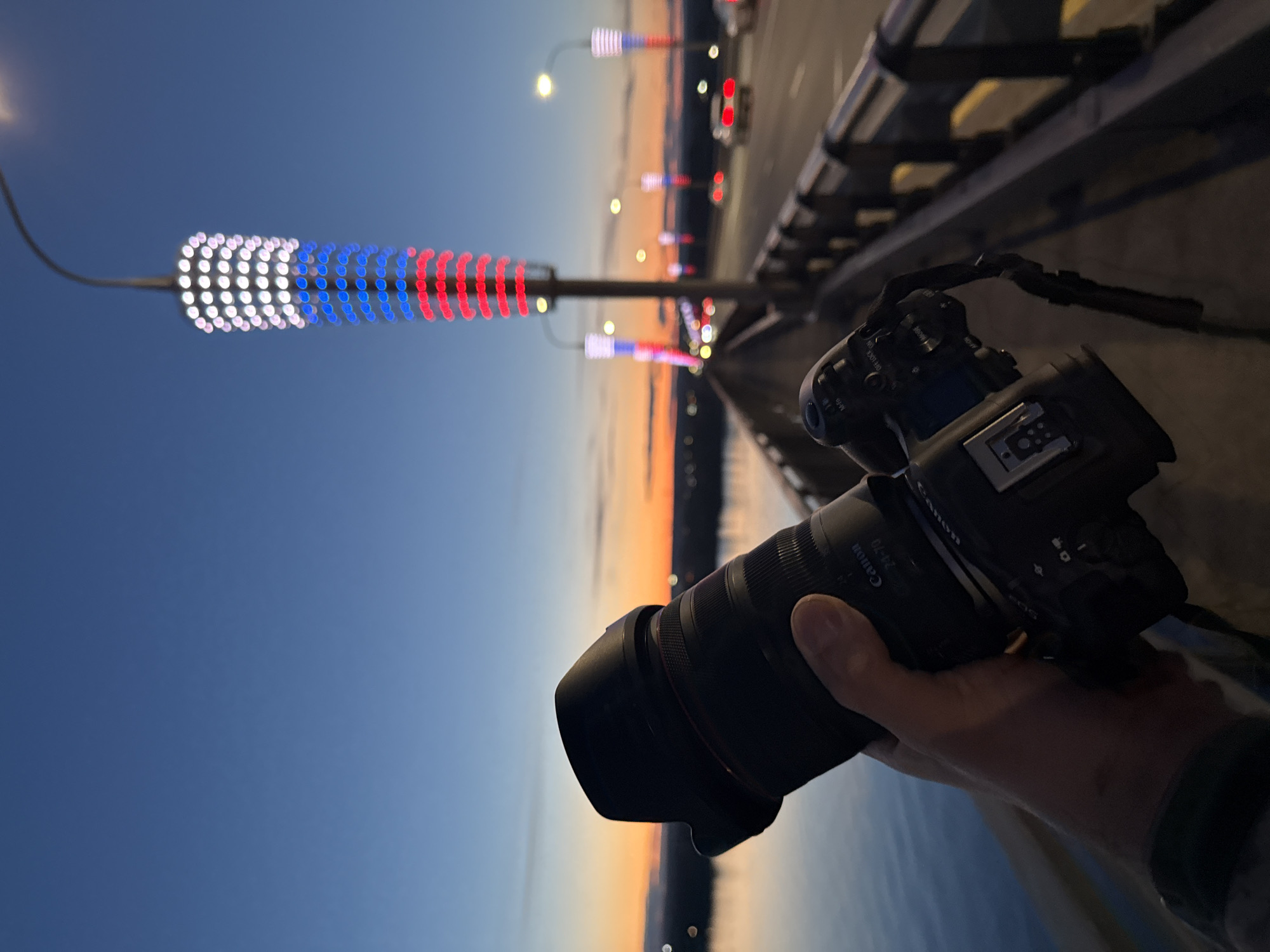 The image captures a handheld view from a bridge in Perm, Russia, at twilight. A Canon EOS R series camera with a 24-70mm lens is in the foreground, held by a person's hand. The cityscape is illuminated by streetlights decorated with white, blue, and red lights, mimicking the colors of the Russian flag. A body of water is visible in the lower portion of the frame, with city lights reflecting on its surface. The sky shows a gradient from blue to orange-yellow, indicating a recent sunset. Traffic and bridge infrastructure, including a guardrail, are visible in the background.