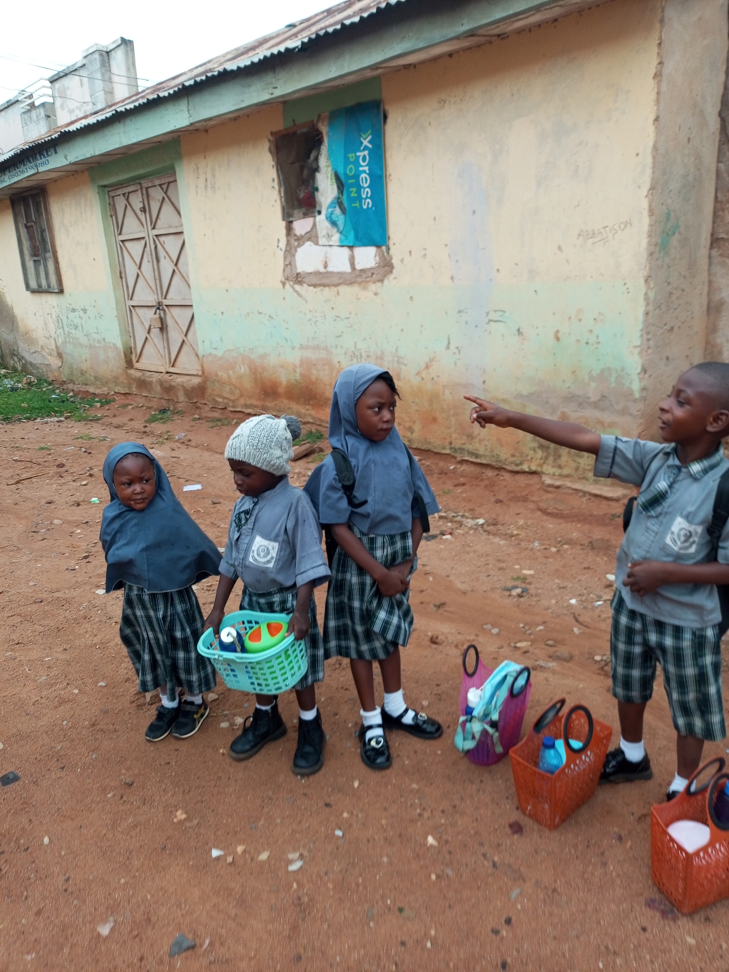 In Tempure, Nigeria, four children are standing on a dirt path in front of a weathered, light-colored building with a green trim. The building has a corrugated metal roof and a locked double door. On the left side of the building, a window with wooden shutters is visible, and on the right, a tattered blue poster with the word "Xpress" is affixed to the wall.
The children, dressed in school uniforms, are the central subjects. The two children on the left are wearing blue hijabs, plaid skirts, and dark shoes. One is holding a small green basket containing what appears to be a toy and some cans. The child in the middle is wearing a knitted gray hat with a pom-pom, a gray shirt, and a plaid skirt. The child on the far right, a boy, is wearing a gray shirt, plaid shorts, and dark shoes. He is pointing to the left, seemingly interacting with the child in the gray hat.
Scattered around the children are various bags and baskets. To the right of the children, there are two orange woven baskets, one containing a blue water bottle and the other empty. A pink mesh bag is also present. The ground is a reddish-brown dirt, with a few scattered stones and debris. The overall scene depicts a moment of everyday life for these schoolchildren, possibly waiting or preparing to go somewhere.