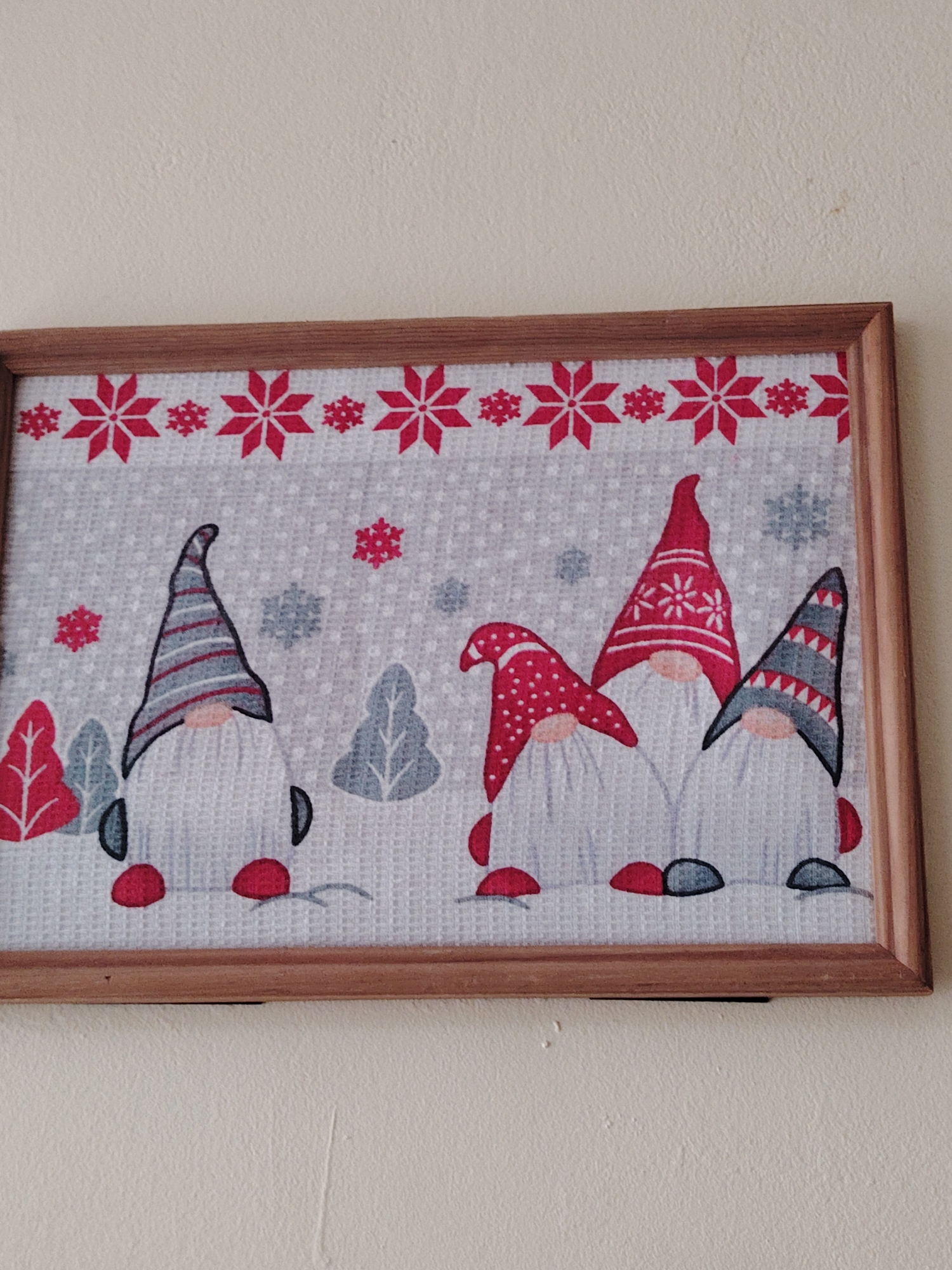 A framed piece of artwork hangs on a cream-colored wall. The artwork depicts three gnomes with tall, patterned hats and red noses. The hats are red and gray with snowflake and stripe patterns. Snowflakes are scattered around the gnomes, and a row of red snowflakes runs along the top of the artwork. The gnomes appear to be standing in a winter scene with stylized trees. The frame is a simple, light-brown wood. The city and location are unknown.