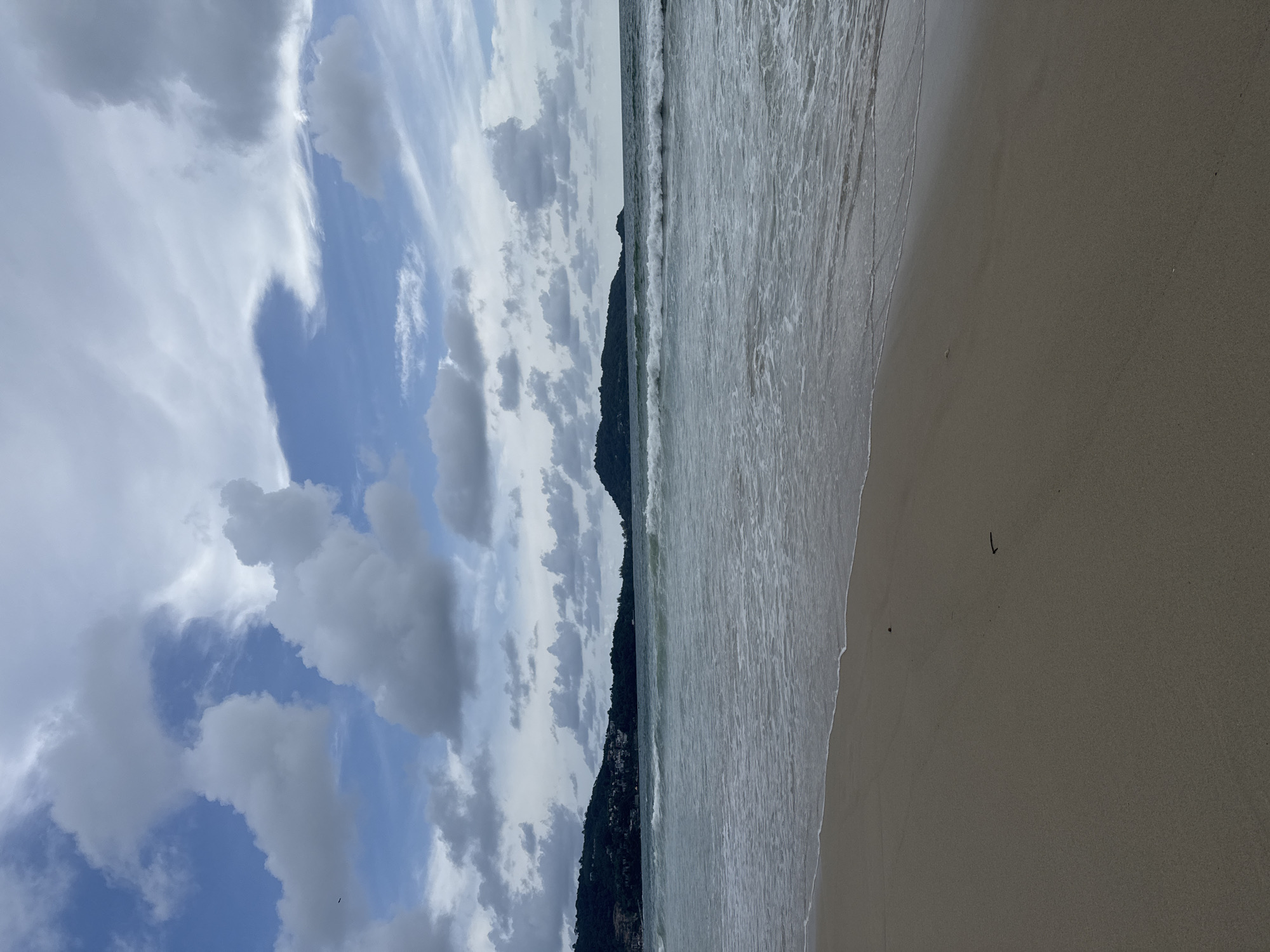 The image captures a tranquil beach scene in Patong, Thailand, under a partly cloudy sky. The foreground is dominated by a sandy beach, with gentle waves lapping at the shore. In the mid-ground, the Andaman Sea stretches out, with small to moderate waves rolling in. The background features lush green hillsides, dotted with buildings, characteristic of the coastal landscape of Patong. A small, rocky island is visible in the distance. The sky is a mix of blue and white clouds, with some wispy formations indicating a dynamic atmosphere. There are no people visible in the frame. The scene is serene and natural, showcasing the beauty of a typical beach day in this popular Thai tourist destination.