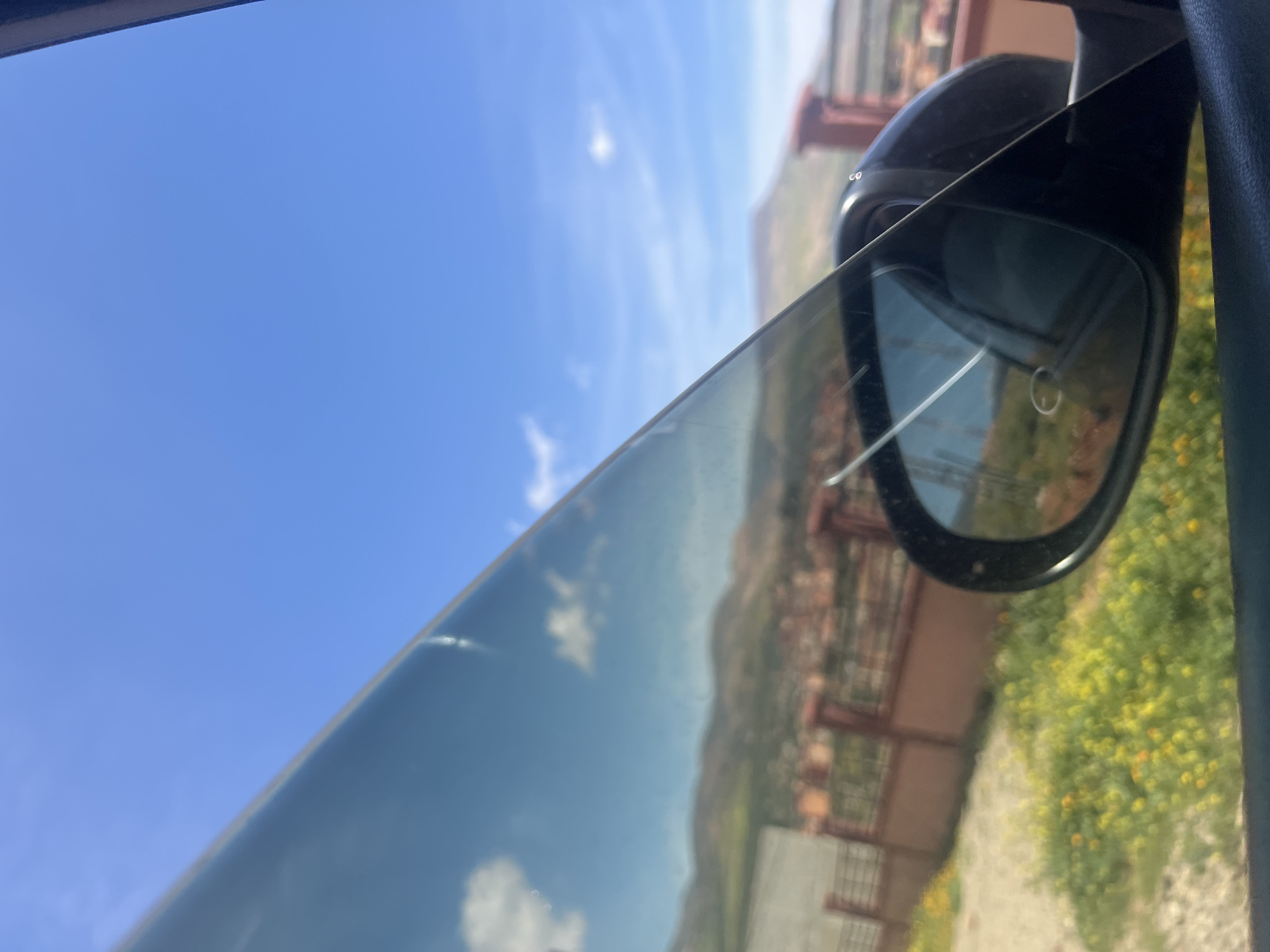 The image shows a view from inside a car, likely traveling through or near the city of Ouaouizert, Morocco. The sky is predominantly blue with a few scattered clouds, visible through the car window. A side mirror is in focus, reflecting the scenery outside, including a blurred view of buildings, possibly houses or shops, and vegetation, likely consisting of plants or grass. The general landscape seems hilly or mountainous, as indicated by the reflected terrain.