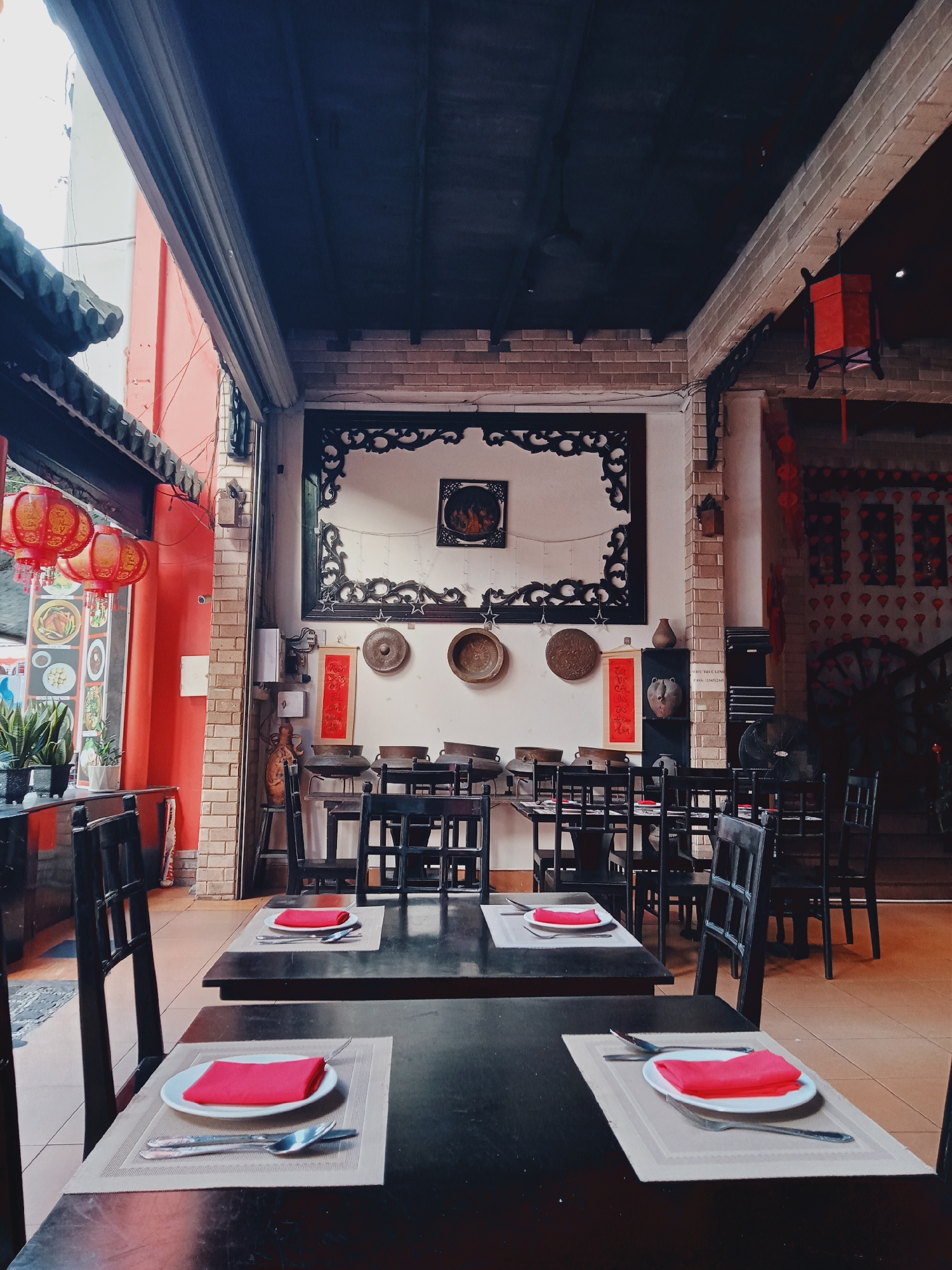 This image depicts the inviting interior of a traditional-style restaurant in Nha Trang, Vietnam, set up for dining during what appears to be daytime. The restaurant is empty of customers or staff, but all tables are meticulously prepared for service.
The scene is characterized by a blend of dark wood furniture and vibrant red and earthy tones in the decor. In the foreground, two dark wooden tables are cleanly set with light beige placemats, white plates, folded bright red cloth napkins, and silverware (forks and spoons). Beyond these, numerous similar dark wooden tables and chairs, featuring simple square-patterned backs, fill the dining area, creating an organized and ready-to-use atmosphere.
Architecturally, the space features exposed brick pillars and dark ceilings with visible beams. The left side of the image opens to an exterior area with a bright red wall, under dark tiled eaves. Several red decorative lanterns with yellow patterns hang outside, and a menu board with images of food is partially visible.
The interior walls are richly decorated with traditional elements. A large, intricate black wooden carving frames a light-colored wall at the back, with a smaller framed artwork hanging within it. Below this, various rustic metallic plates or gongs, a red scroll with Asian script, and large dark pots serve as decorative accents. Another section of the wall on the right side features numerous smaller red decorations and a large, dark, circular lattice design, possibly representing a cartwheel. A black electric fan is visible on the floor in the background.
A notable detail is a white sticker on a brick pillar, clearly displaying Wi-Fi credentials: "WIFI: TRUC LING" and "PASS: 123412345." The restaurant's overall aesthetic combines functionality with a strong cultural identity, suggesting a warm and welcoming dining experience.