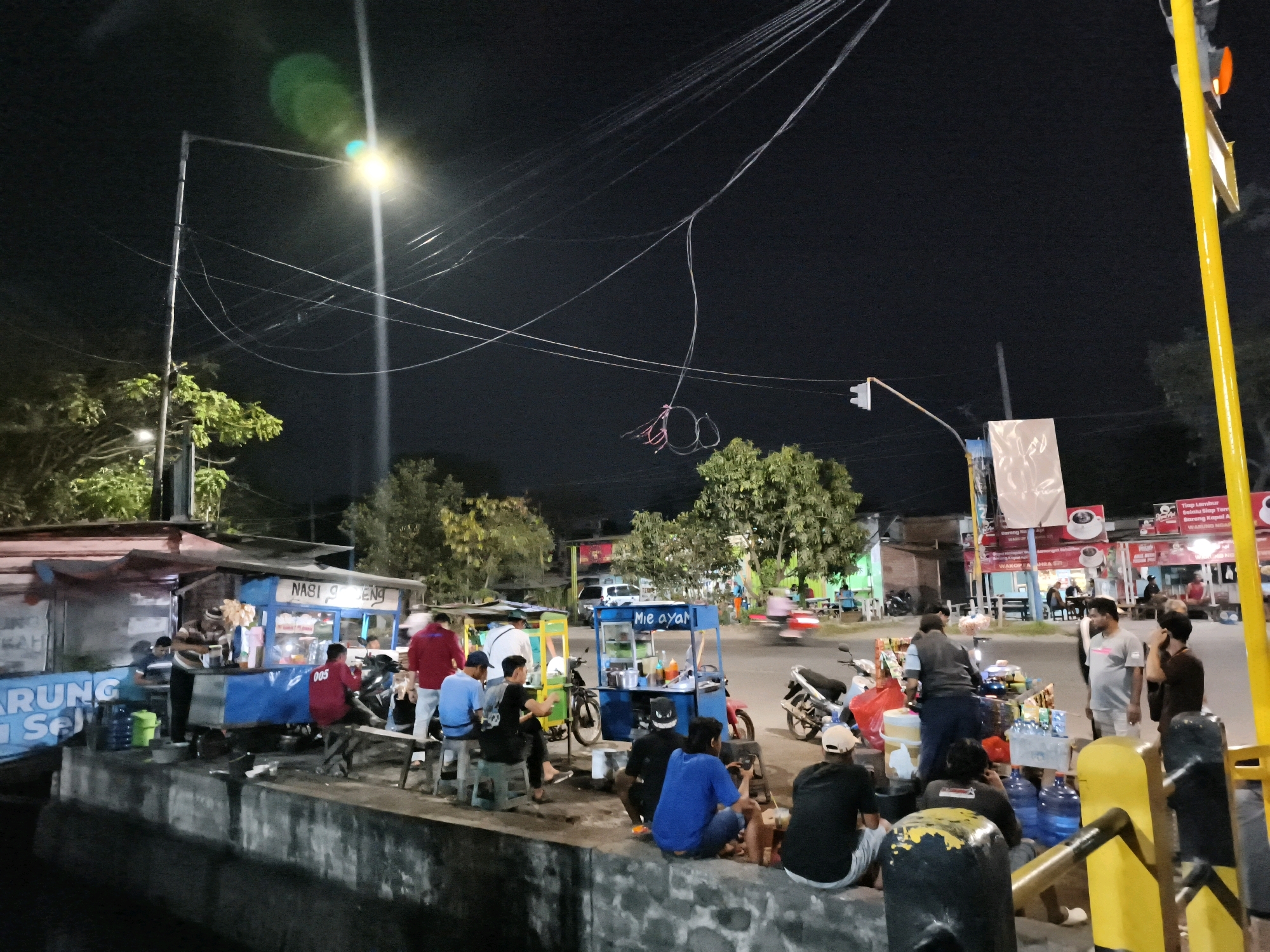 The image depicts a lively night scene in Desa Ponokawan, Indonesia, likely a bustling street food area. Several food stalls are set up along a raised concrete barrier, with people gathered around them. One stall prominently displays "Nasi Goreng" and another "Mie Ayam," indicating popular local dishes. People are seated on stools and the ground, consuming food and drinks. There are several motorcycles parked nearby, suggesting that transportation is a common mode for patrons. Streetlights and the illuminated signs of the stalls provide the primary light sources, creating a warm and inviting atmosphere. The background shows trees, buildings, and a traffic light, further establishing the urban setting. The overall impression is one of a vibrant community gathering spot, active during the evening hours for dining and socializing.