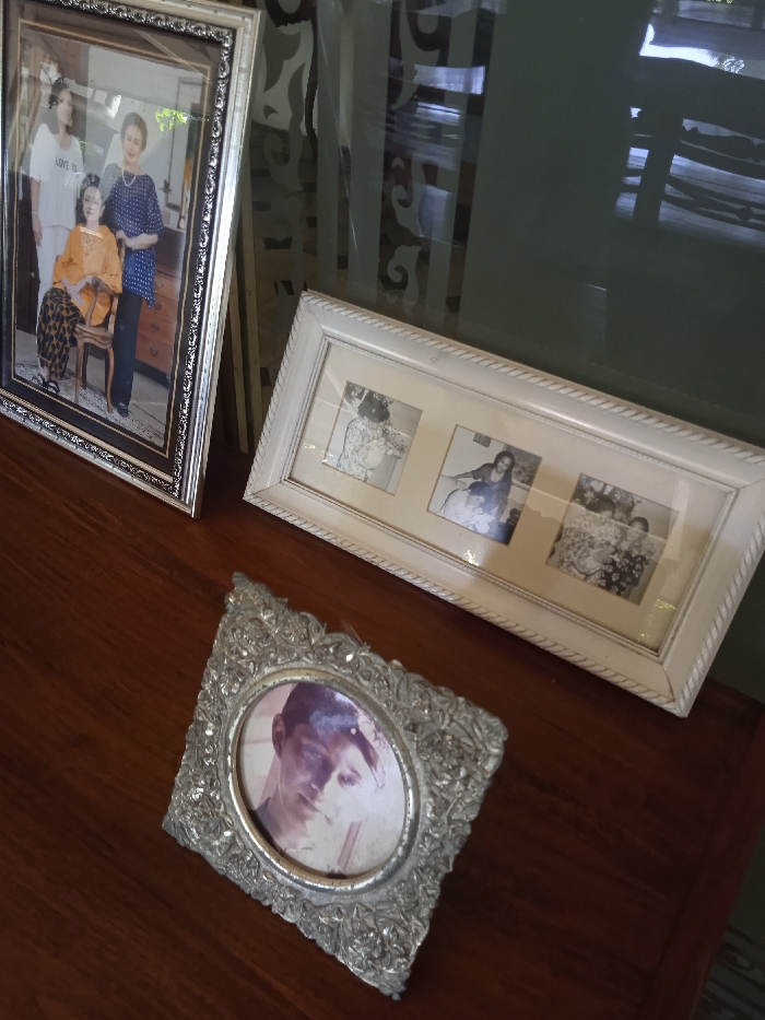 The image shows a wooden table top, with three picture frames. One is an ornately framed portrait of a man with dark hair, and the other two are rectangular, white-painted frames with photos inside. The frames are arranged in a triangle shape, with the smaller, ornate frame at the bottom. The two white frames contain black and white photographs of a woman standing in a garden setting, two women standing in a more formal setting, and two women and a child in an outdoor, casual setting. The image was taken in the city of Surakarta, Indonesia, at the location with the coordinates -7.566 latitude, 110.823 longitude.
