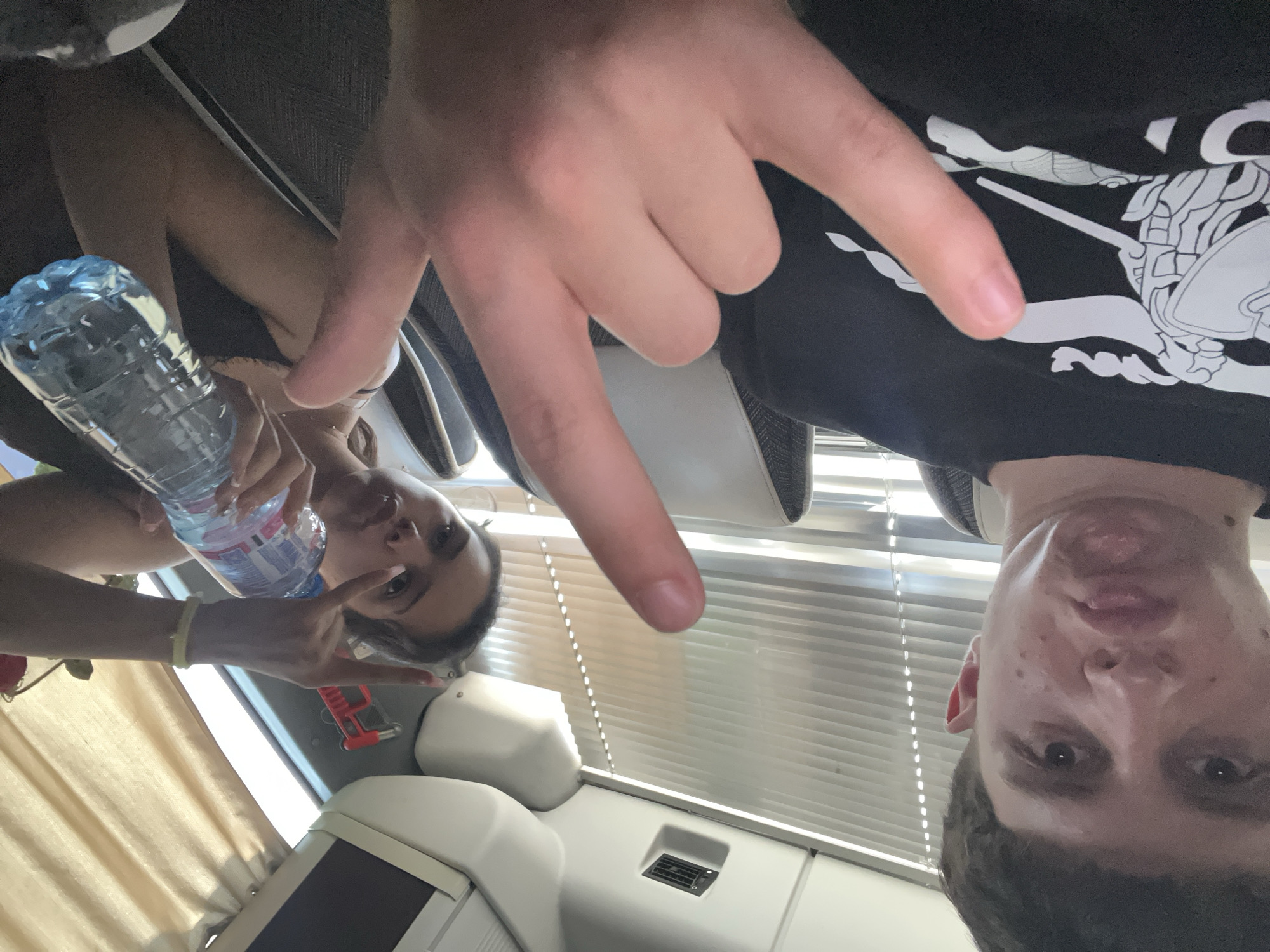 Two individuals are depicted in a selfie-style image. A female, holding a water bottle, is visible in the left portion of the frame making a hand gesture. To her right is a red toy car. A male is to the right, wearing a black t-shirt with a white design, also making a hand gesture. The background suggests an interior setting with window blinds visible. The location cannot be identified from the information given.