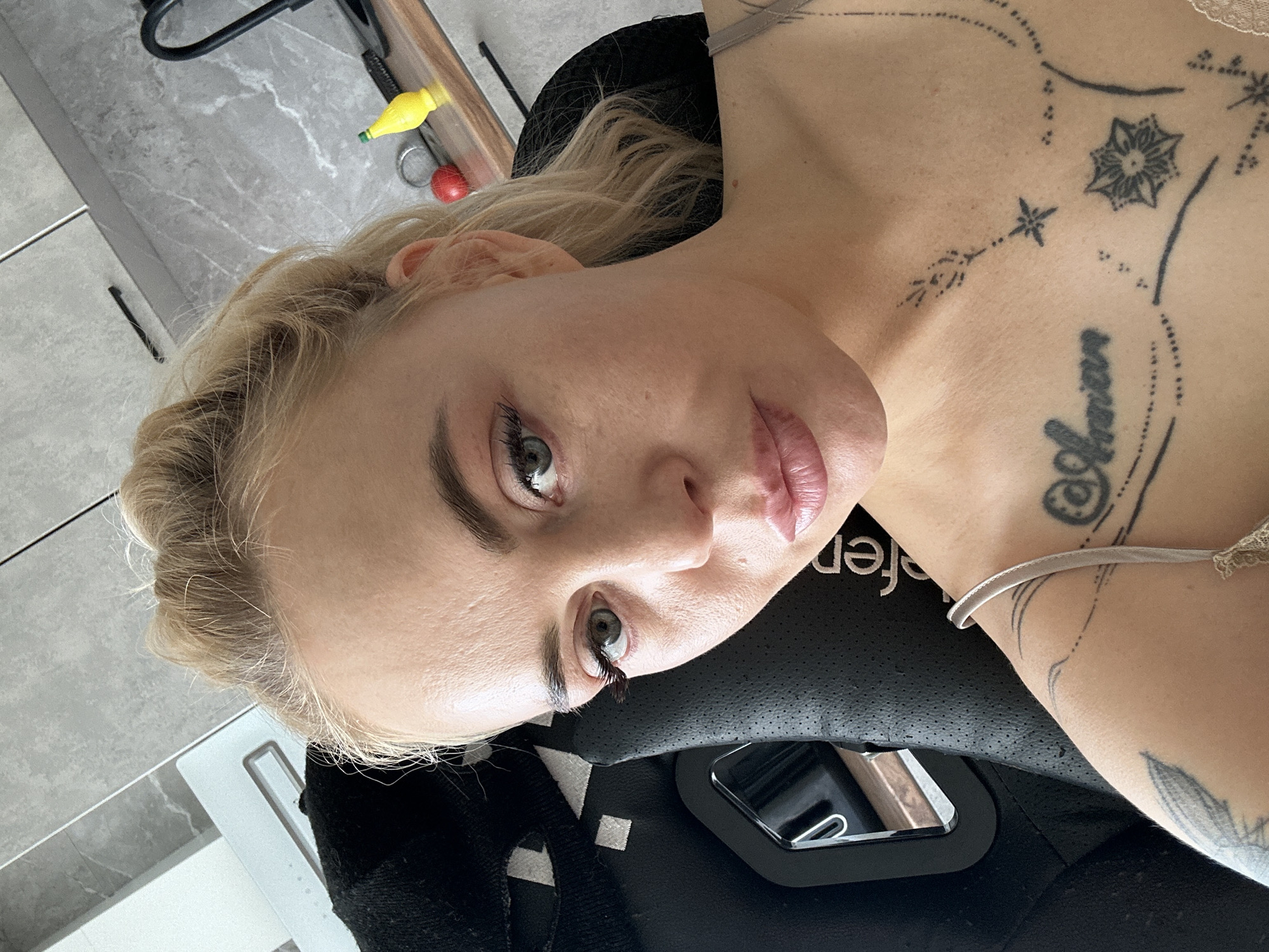 This close-up selfie captures an adult female with blonde hair styled loosely, looking directly at the camera. Her light-colored eyes are visible, along with defined eyebrows and makeup on her eyes and lips. Multiple black ink tattoos adorn her upper body: a script design, possibly reading "Annar," on her central chest, and intricate dot-work patterns featuring stars and a mandala-like flower on her right chest and shoulder. A partial tattoo resembling a feather or leaf is visible on her left shoulder. She is wearing a light-colored, thin-strapped top. The subject is seated in a black gaming chair with perforated material, the word "fer" or "er" partially visible on its side, and a white geometric pattern on its backrest. The background reveals a modern interior with grey, marble-patterned cabinetry featuring black handles. A wooden countertop on the right displays a small red spherical object, a shiny metal ring, and a yellow lemon-shaped bottle with a green cap. A white rectangular object, possibly an appliance or control panel, is visible on the upper left.