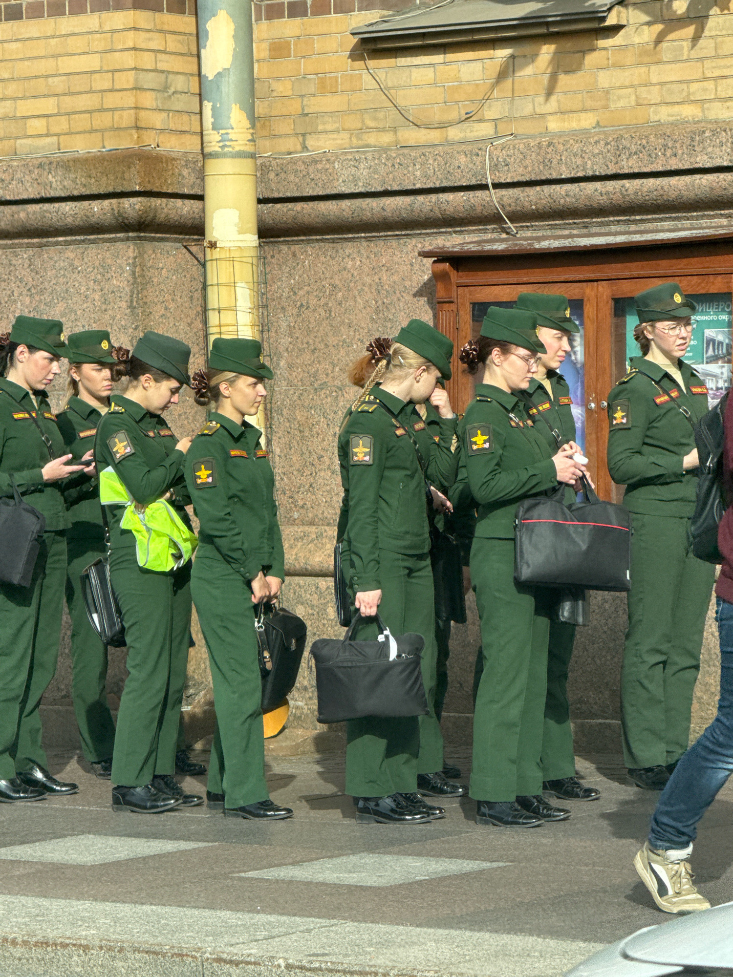 A group of women in green military uniforms stand in a line on a sidewalk in Saint Petersburg, Russia. The women are wearing peaked caps and have their hair styled in buns. They are all carrying black bags, and some are holding phones. The background consists of a brick building with a large wooden door and a stone facade. A metal pole with peeling paint is visible on the left side of the image. The women appear to be waiting or marching, and the overall scene suggests a formal or ceremonial occasion.