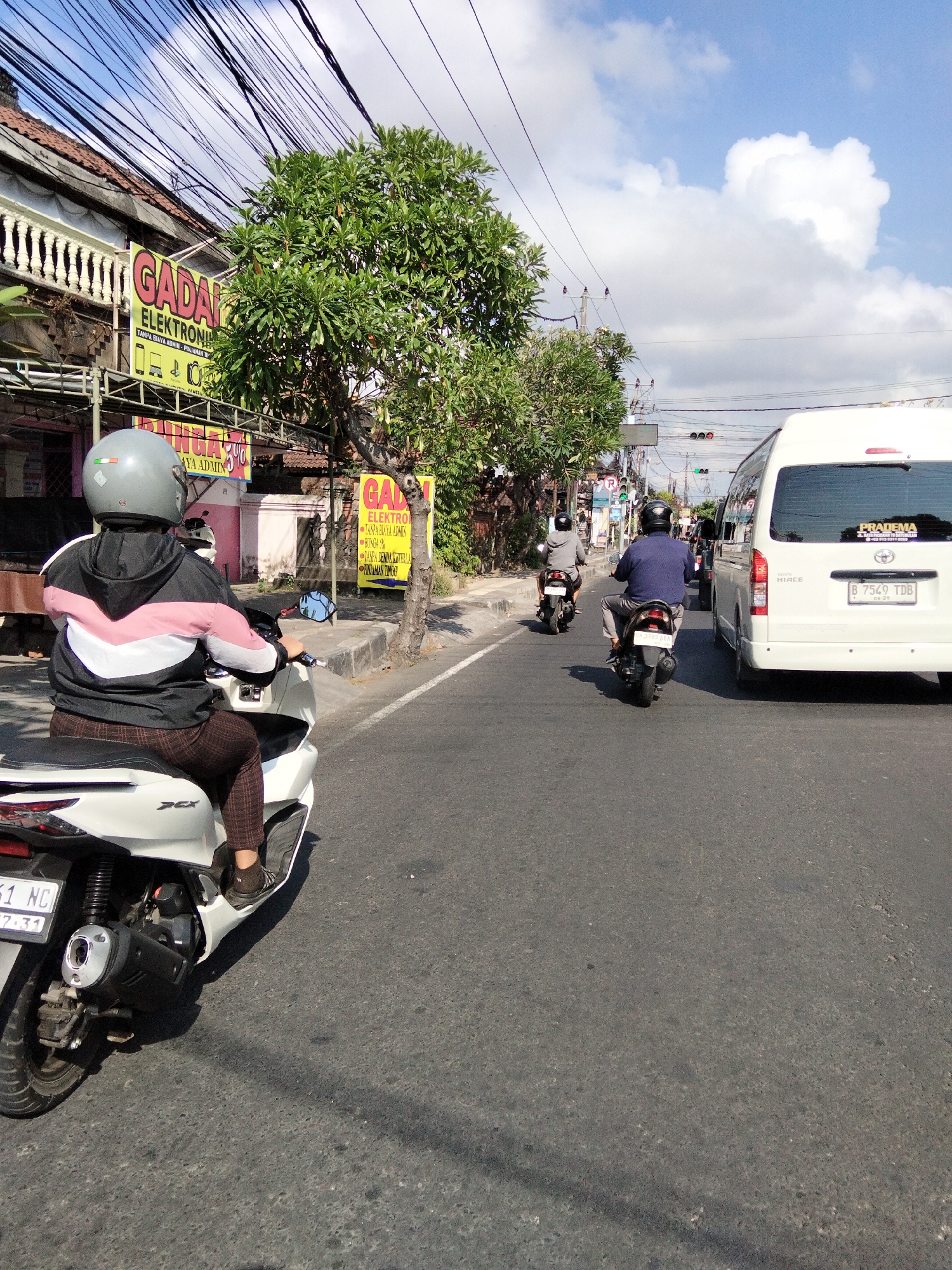 The image captures a street scene in Denpasar, Indonesia, on a sunny day with scattered clouds. The viewpoint is from behind a person riding a white scooter, looking forward down a two-lane road. The scooter has a license plate visible, indicating it is registered in Bali. The rider wears a grey helmet and a jacket with a chevron pattern in pink, white, and black, paired with brown checkered pants.
On the left side of the road, a building with a white decorative railing is visible, adorned with a yellow sign advertising "GADA ELEKTRONIK" and another sign below it mentioning "BUNGA 0%". A large green tree with sparse foliage is situated near the building, casting a shadow on the road. Several utility wires are strung overhead, crossing the frame.
Further down the road, another scooter rider, wearing a dark helmet and a navy blue shirt, is also moving in the same direction. A white van, identified as a Toyota Hiace, is in the right lane, ahead of the scooter riders. The van has a license plate with the characters "B 7549 TDB" and a sticker on the rear window reading "PRADEMA". Traffic lights are visible in the distance, indicating an intersection. The road surface is asphalt, with some wear and tear. The overall atmosphere suggests a typical urban street with ongoing traffic.