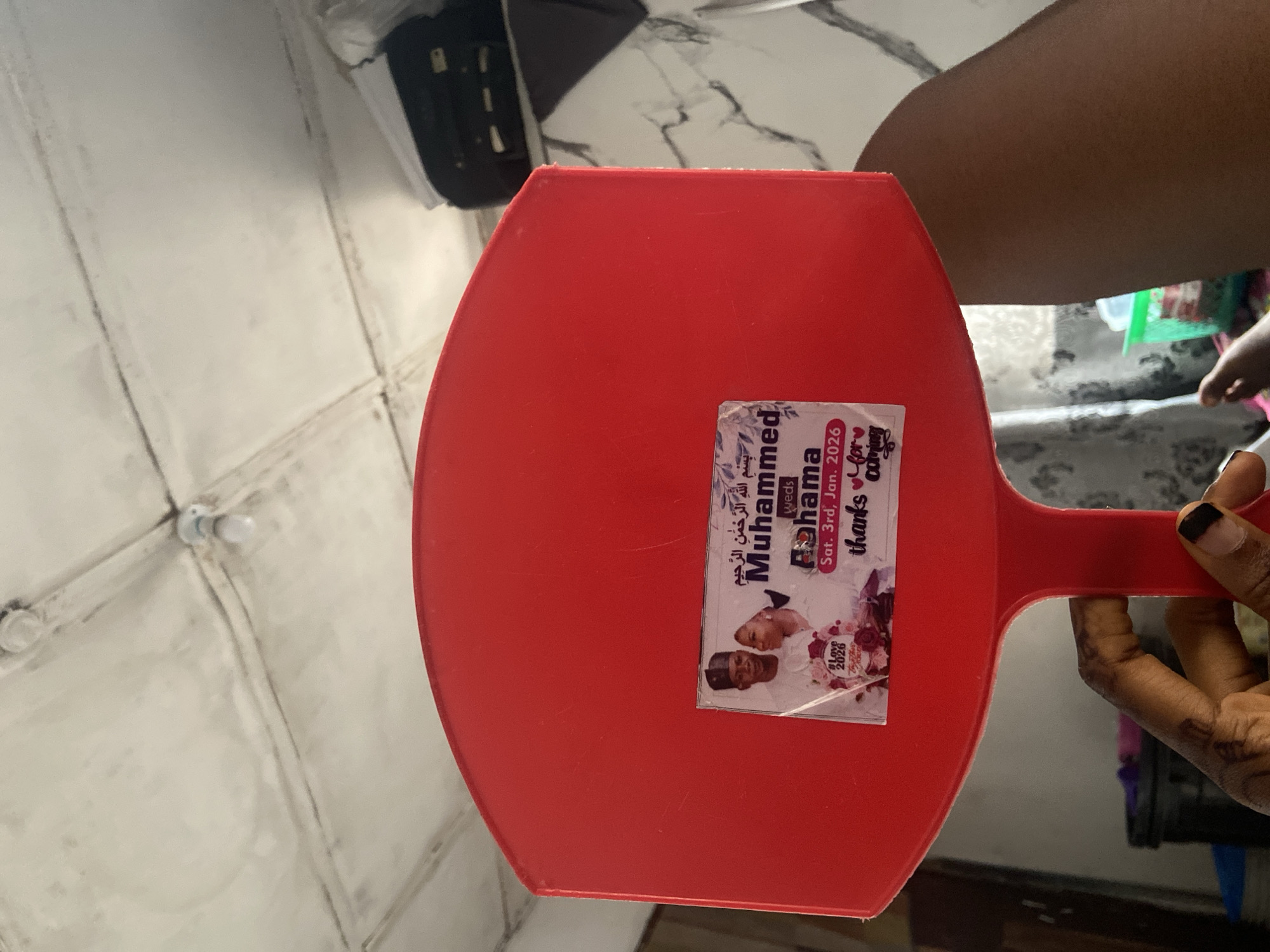 A person is holding a red dustpan with a sticker on it. The sticker has an image of a couple, presumably getting married, and the text "بِسْمِ اللَّهِ الرَّحْمَنِ الرَّحِيمِ Muhammed weds Pohama Sat. 3rd, Jan. 2026 thanks for coming". The background includes a white tile wall and other indistinct objects. The location is unknown, so it is not possible to say if it is in Lokoja, Nigeria.