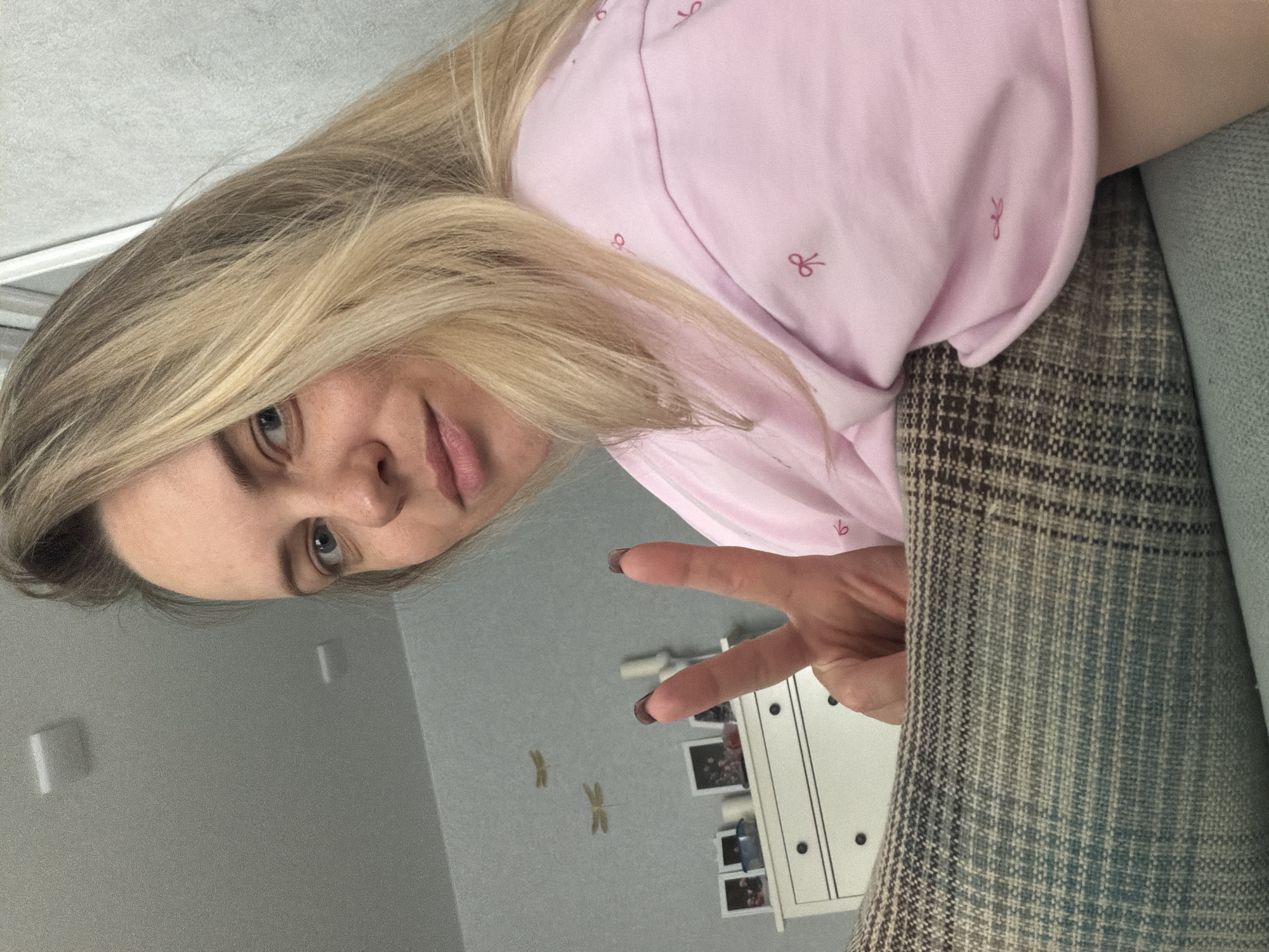 A woman with blonde hair and blue eyes is visible from the chest up, wearing a pink shirt with bow patterns. She is making a peace sign with her fingers. The background features a gray wall with dragonfly decorations, a white cabinet with pictures, and a patterned gray and brown couch. There is no information in the image to specify the location in the city of Votkinsk, Russia.