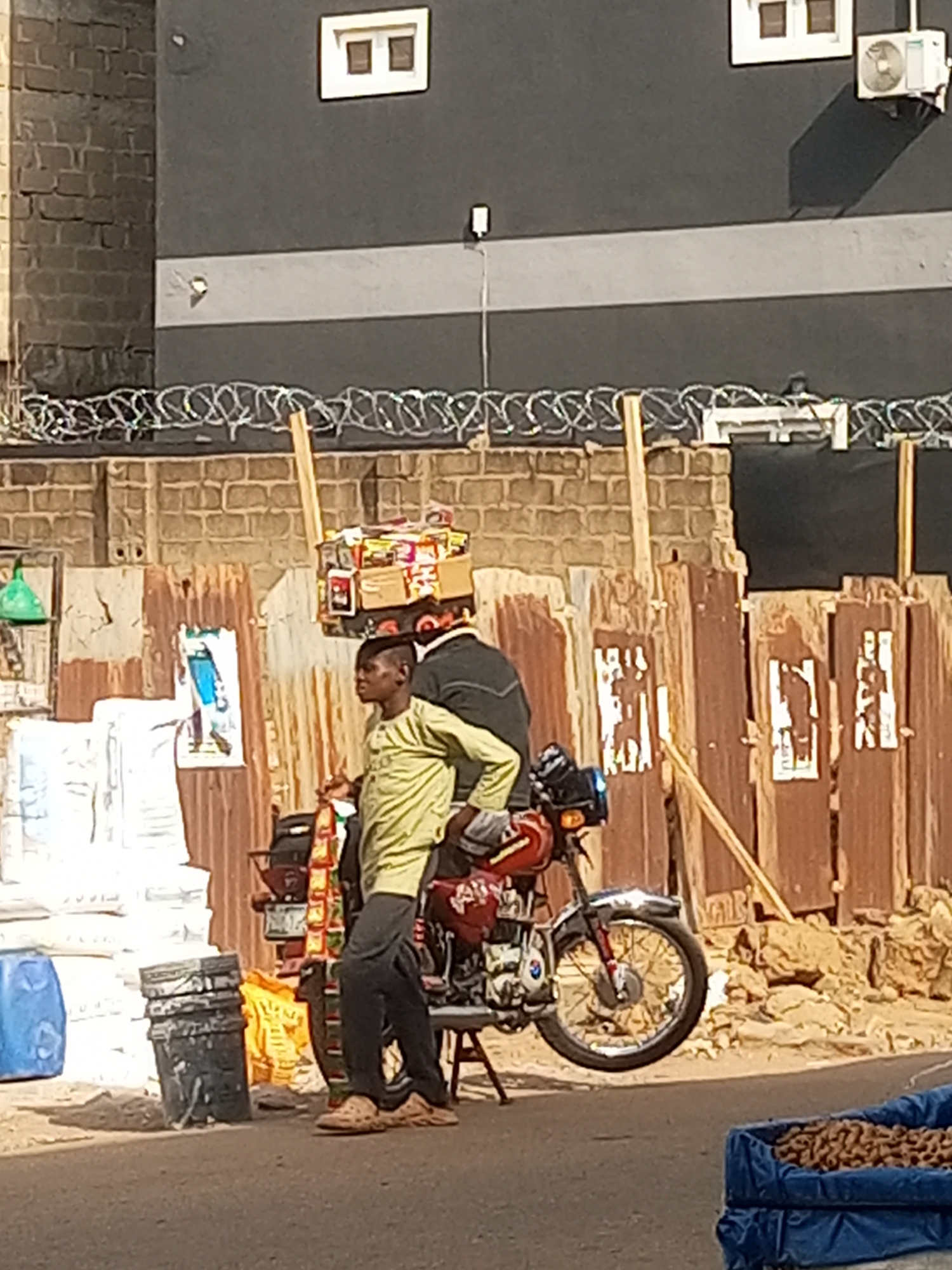 In Maiburiji, Nigeria, a man stands next to a parked red and silver motorcycle on a paved street. He wears a light green, long-sleeved shirt and dark pants. On his head is a stack of boxes and packages. A second person in a dark jacket is on the motorcycle. To the left of the motorcycle are stacked white bags and black buckets. Behind them is a fence made of corrugated metal sheets with faded posters. Above the fence, there is a brick wall with barbed wire. In the background, a gray building with windows and an air conditioning unit is visible. To the right of the motorcycle, the ground is covered in dirt and rocks. In the foreground, a blue container is filled with brown contents.