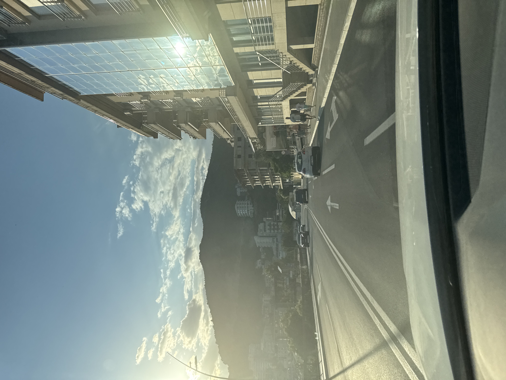 The image depicts a street scene in Boreti, Montenegro, at sunset. The perspective is from inside a vehicle, looking forward. A multi-lane road with white lane markings and directional arrows occupies the foreground. To the right, a modern building with a facade of reflective glass windows and multiple balconies rises, with a staircase leading up to the entrance. Several vehicles are visible on the road, including a white car and at least two darker cars. A few pedestrians are walking on the sidewalk in front of the building. In the background, a hazy, sun-drenched mountain range is visible, dotted with buildings of varying architectural styles. The sky is a clear blue with scattered white clouds, and the setting sun casts a warm, golden light over the entire scene.