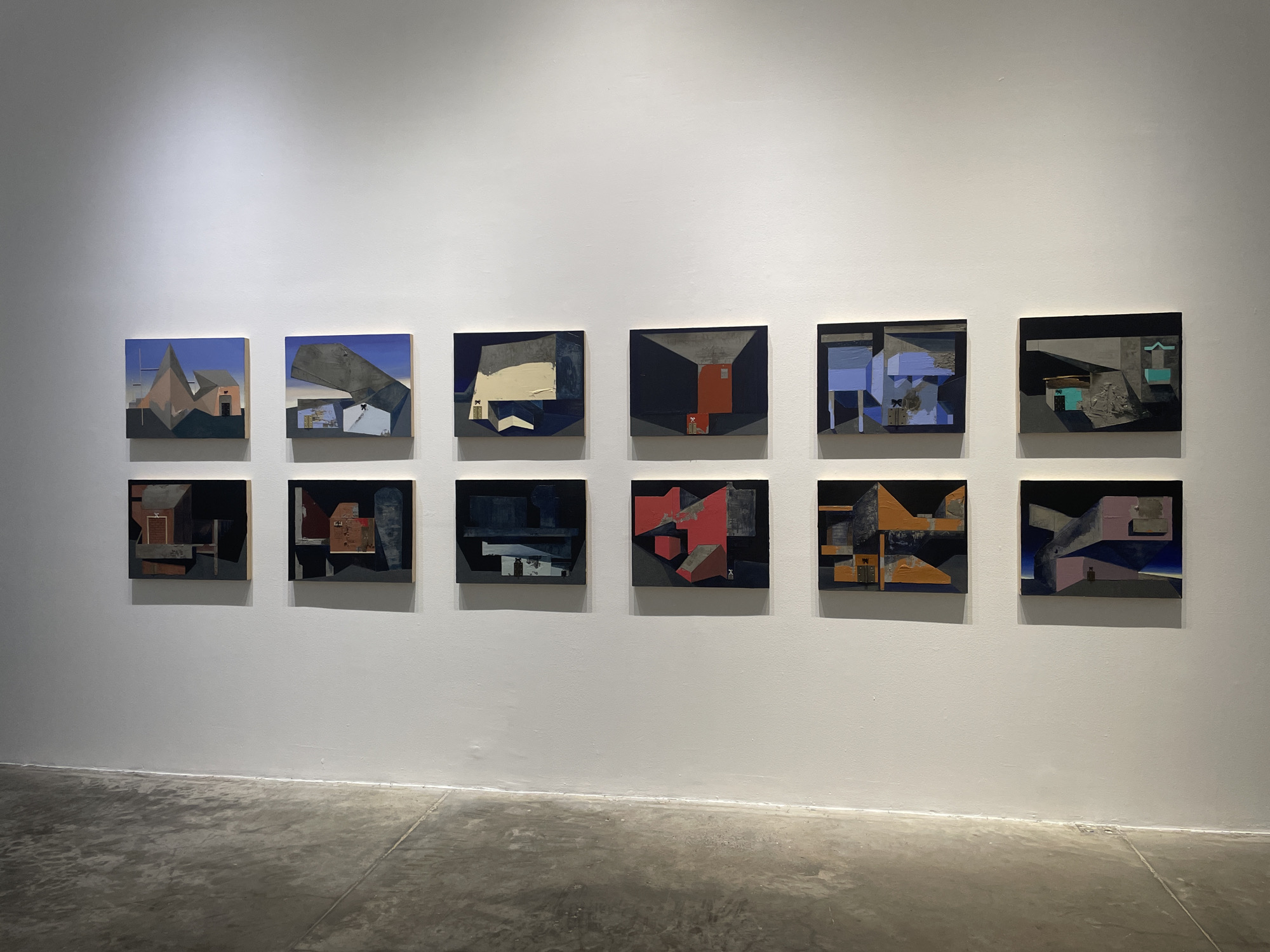The image shows a white-walled room, likely a gallery, featuring a display of ten small, square, abstract paintings hung in two rows. The paintings have a dark, muted color palette with geometric shapes. The foreground displays a light-gray concrete floor. There are no people in the image. The location may be in Mexico City, Mexico.