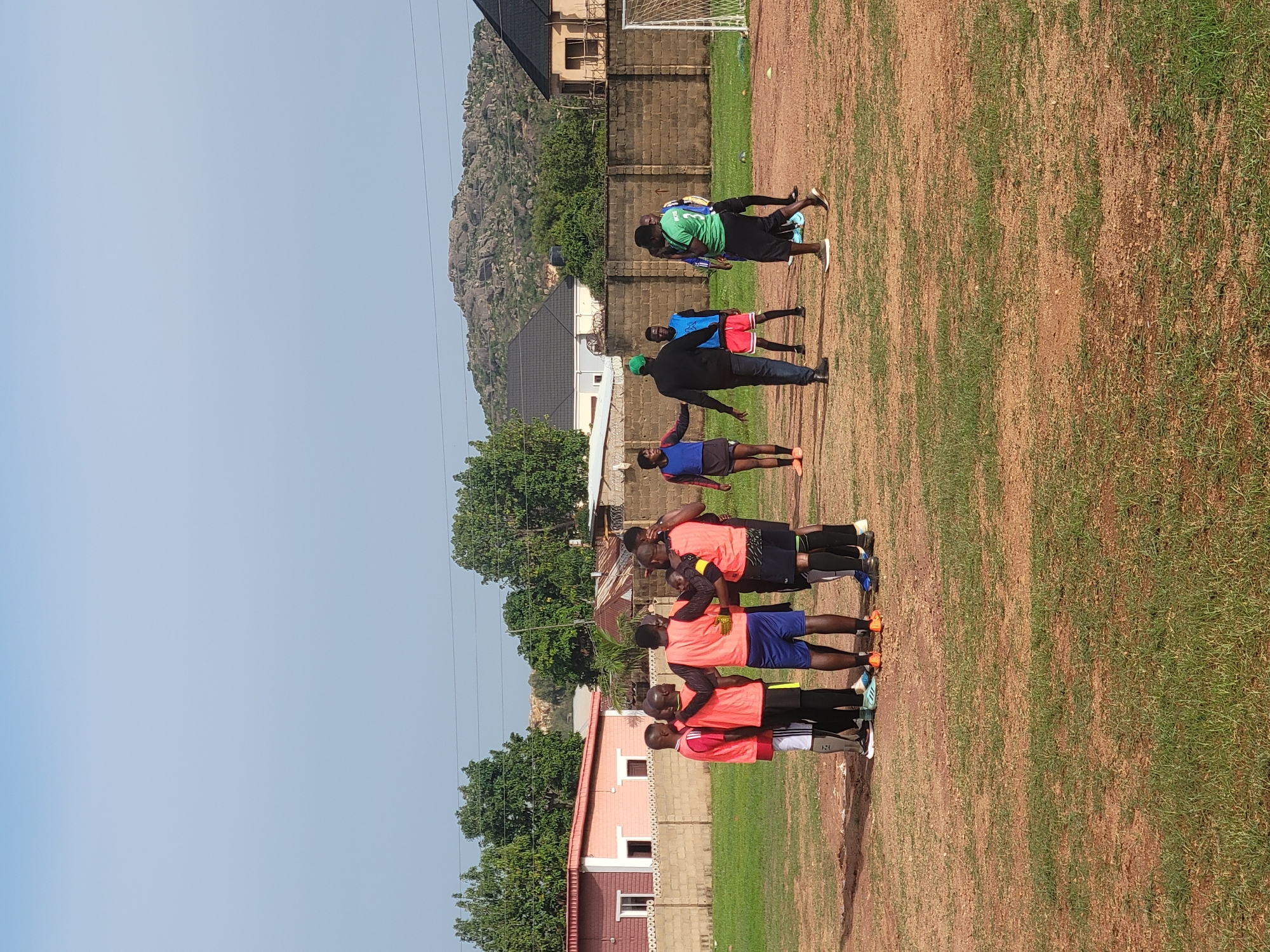 The image depicts a group of people, likely a soccer team and coach, outdoors on a grassy field, possibly a training ground, in the city of Jos, Nigeria. The environment includes buildings, trees, and a rocky outcrop in the background.
People:
A group of individuals are present, wearing soccer training attire, lined up together.
A coach is present, guiding the group.
There are about 10 people in the photo, including the coach and the players.
The players are wearing different uniform colors. Some have orange vests and blue shorts, others have green tops.
Objects:
Soccer training equipment.
Buildings, possibly residential or institutional.
Trees and other vegetation.
A goalpost structure is visible in the background.
Electrical power lines are overhead.
Actions and Interactions:
The group is standing together, likely receiving instructions or preparing for a training session.
Environment:
Outdoor setting with a grassy field and surrounding buildings.
The sky is clear and sunny.
Jos, Nigeria, is suggested by the presence of rocky hills and the likely style of architecture.