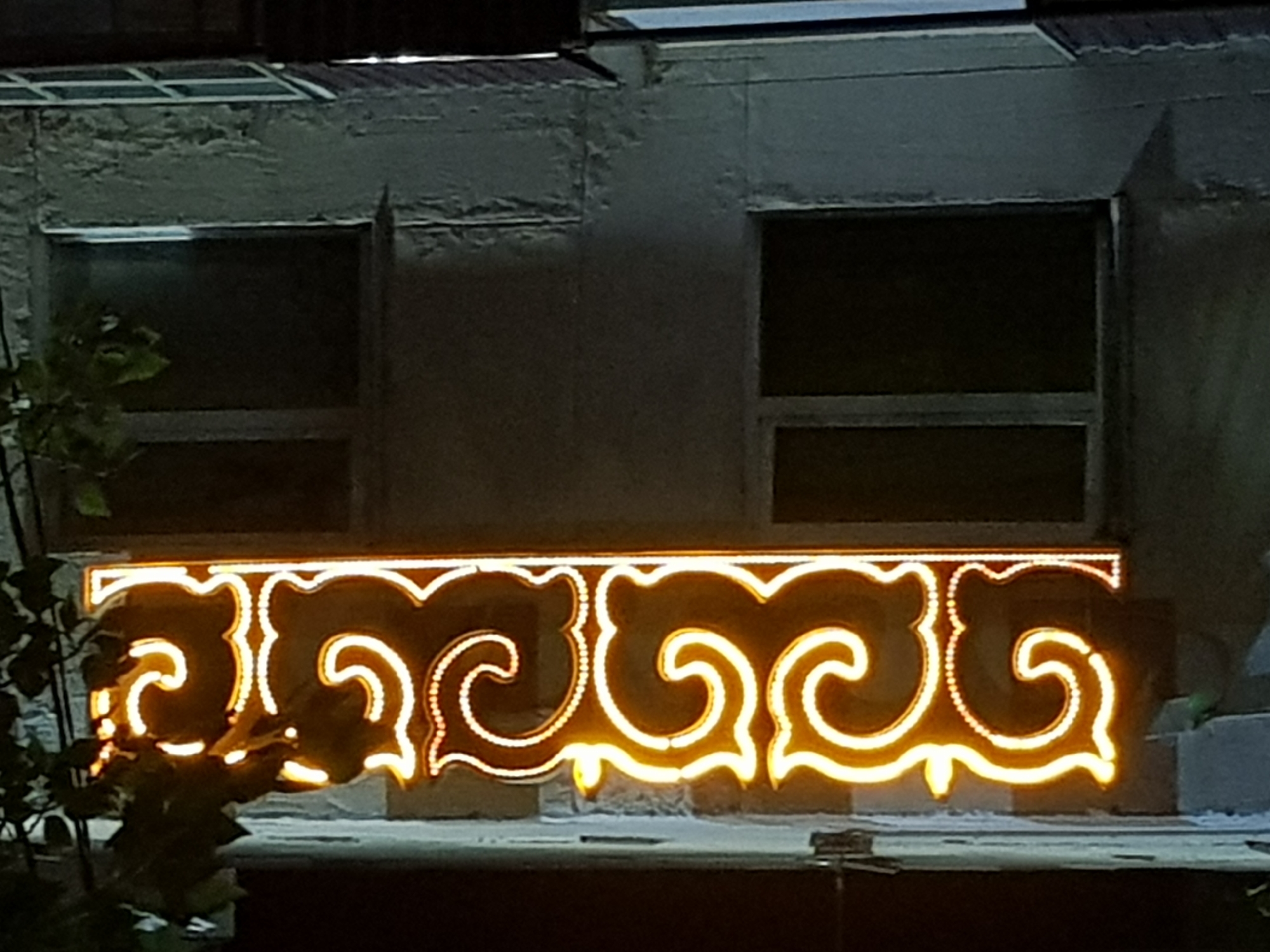 An illuminated neon sign with ornate yellow swirls and dots adorns the side of a weathered concrete building. The sign is positioned vertically and displays a repeating pattern of curved shapes. Adjacent to the sign are two dark, recessed windows on the upper and lower floors of the building. The building's exterior shows signs of age, with peeling paint and a rough texture. Dark green leaves from an unseen tree overhang the top portion of the neon sign. The overall scene is illuminated by the bright neon light, with the surrounding environment appearing dark, suggesting it is nighttime. The location is identified as Kurchatov, Kazakhstan.