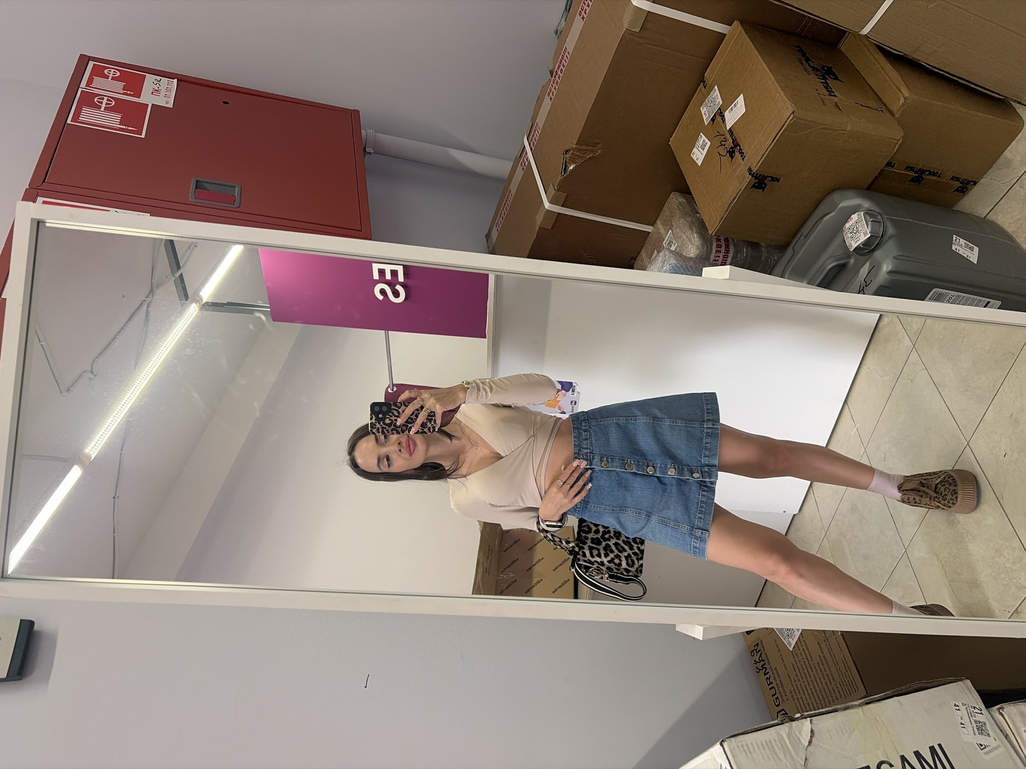 A light-skinned female with long dark hair stands in a full-length, white-framed mirror, taking a selfie with a smartphone. She is wearing a beige long-sleeved wrap top, a blue button-front denim mini-skirt, light pink socks, and leopard-print platform shoes. Her accessories include multiple gold rings, a gold necklace with a cross pendant, a leopard-print smartphone case, and a leopard-print handbag with a black and white striped strap resting on her left forearm. She is looking towards the phone with a slight smile.
The environment depicts a utilitarian commercial or storage space. The walls are painted a light gray. To the right of the mirror, a red fire hose cabinet is mounted on the wall, marked with "ПК-52" and "тел. 01.101.112" (standard Russian fire safety designations). The floor is covered with light-colored square tiles.
Around the mirror and reflected within it are various stacked cardboard boxes, indicating a storage or backroom area. One large box prominently displays the Russian text "ОСТОРОЖНО! ХРУПКОЕ!" (Caution! Fragile!). Other boxes bear the branding "TOOLTECH" and various shipping labels. Another visible box, for "GURMAN" brand refined sunflower oil, includes detailed Russian text indicating its origin: "ООО «БЛОКНОТ»", with legal and actual addresses in Volkhov, Leningrad Oblast, Russia (187401, Leningradskaya obl., g. Volkhov, ul. Voroshilova, d.42, k. A and 187402, Leningradskaya obl., g. Volkhov, ul. Kirova, d.37). A gray 20-liter plastic canister is also visible among the stacked boxes. The reflection in the mirror shows overhead fluorescent strip lighting and a purple sign with the reversed characters "ES" (likely "23" when viewed from the front).
The scene is a generic commercial backroom or storage facility in Russia, identifiable by the Cyrillic text on packaging and fire safety equipment. Specific visual or textual identifiers within the image that definitively locate the scene within the city of Saint Petersburg are absent; however, the environment is consistent with commercial spaces found throughout Russian cities.