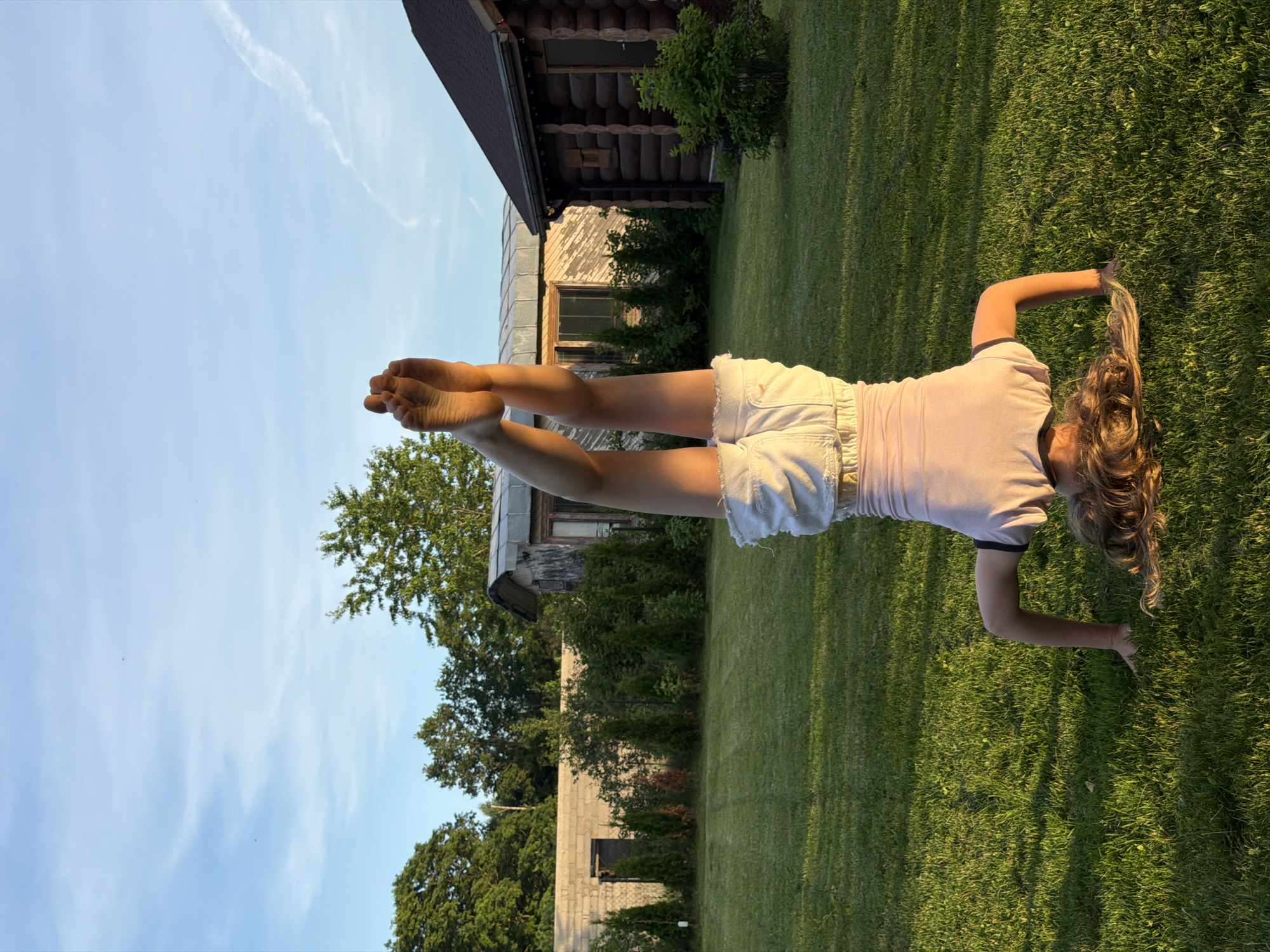 In the city of Dreiliņi, Latvia, a young person is performing a headstand on a lush green lawn. They are wearing a light pink t-shirt with a dark trim and white denim shorts. Their legs are extended upwards, and their bare feet are visible against a bright blue sky with wispy clouds. The background features a rustic wooden cabin with a dark brown roof, a small building with weathered siding, a low concrete wall, and several evergreen trees. The setting appears to be a backyard or rural property, with the warm glow of sunset casting long shadows across the grass.