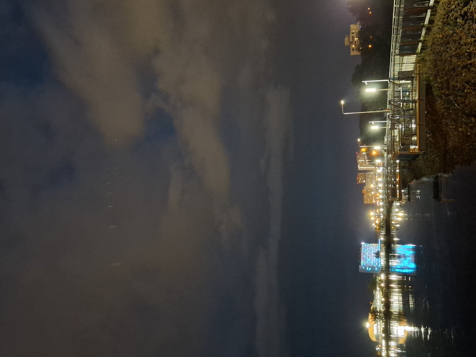 The image depicts a nighttime scene along a waterfront in Tyumen, Russia. In the foreground, a dark body of water reflects the lights of the cityscape. A wooden and metal pier extends into the water, featuring lampposts and seating areas. Beyond the pier, a cityscape is visible with several illuminated buildings, including a prominent building with bright blue lighting. The sky is dark and cloudy, with some lights reflecting off the cloud cover. The overall atmosphere is calm and serene.