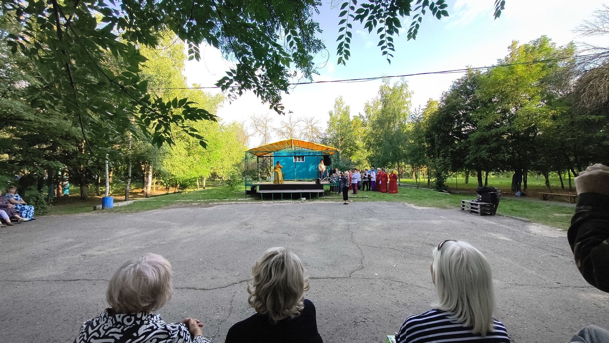 The image shows an outdoor performance in a park, possibly in Mikhaylovka, Russia, given the context. In the foreground, three individuals with their backs to the camera are seated on the ground, facing a stage. The stage, a raised platform with a yellow canopy, hosts a performance. On the stage, a woman in a yellow dress is performing. Several other people, some in red traditional clothing, are gathered on or near the stage, appearing to be part of the performance or an audience. The park environment is characterized by lush green trees and a clear sky. The ground is a paved area, suggesting a dedicated performance space within the park.