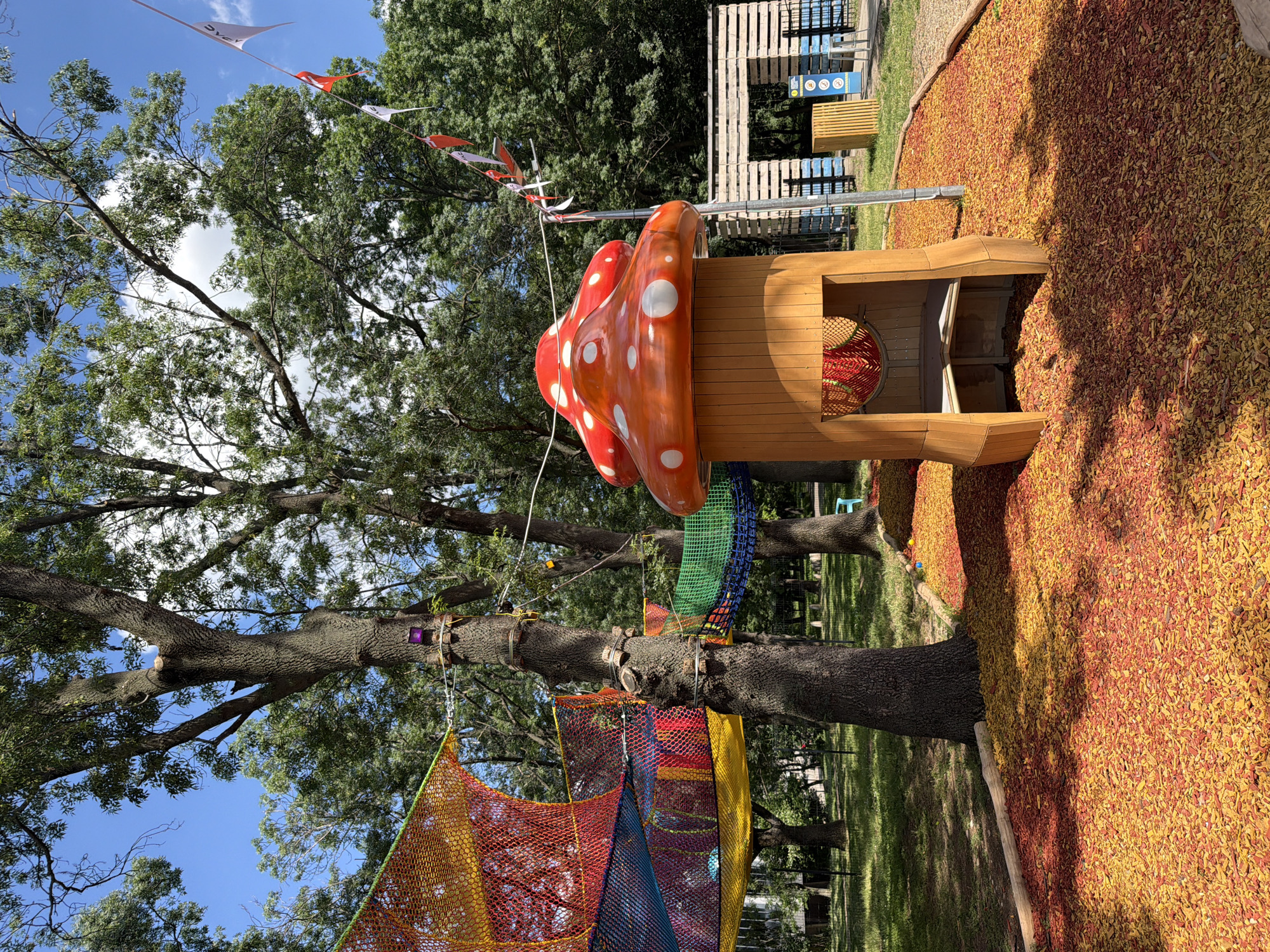 The image depicts an outdoor playground in Sofia, Bulgaria, bathed in daylight under a clear blue sky with scattered white clouds. The playground features a whimsical, mushroom-themed play structure with a red cap adorned with white polka dots, and a wooden base. Attached to this structure and a large tree are colorful nets, forming a climbing or play area. The ground is covered in a thick layer of red and yellow wood chips, providing a soft landing surface. In the background, a wooden structure with multiple levels and benches suggests a seating or viewing area. Flags or banners, some triangular and white with red accents and a logo that is illegible, are strung across the scene, adding a festive touch. The overall atmosphere is cheerful and inviting, suitable for children's recreation. The shadows cast by the trees indicate it is likely midday.