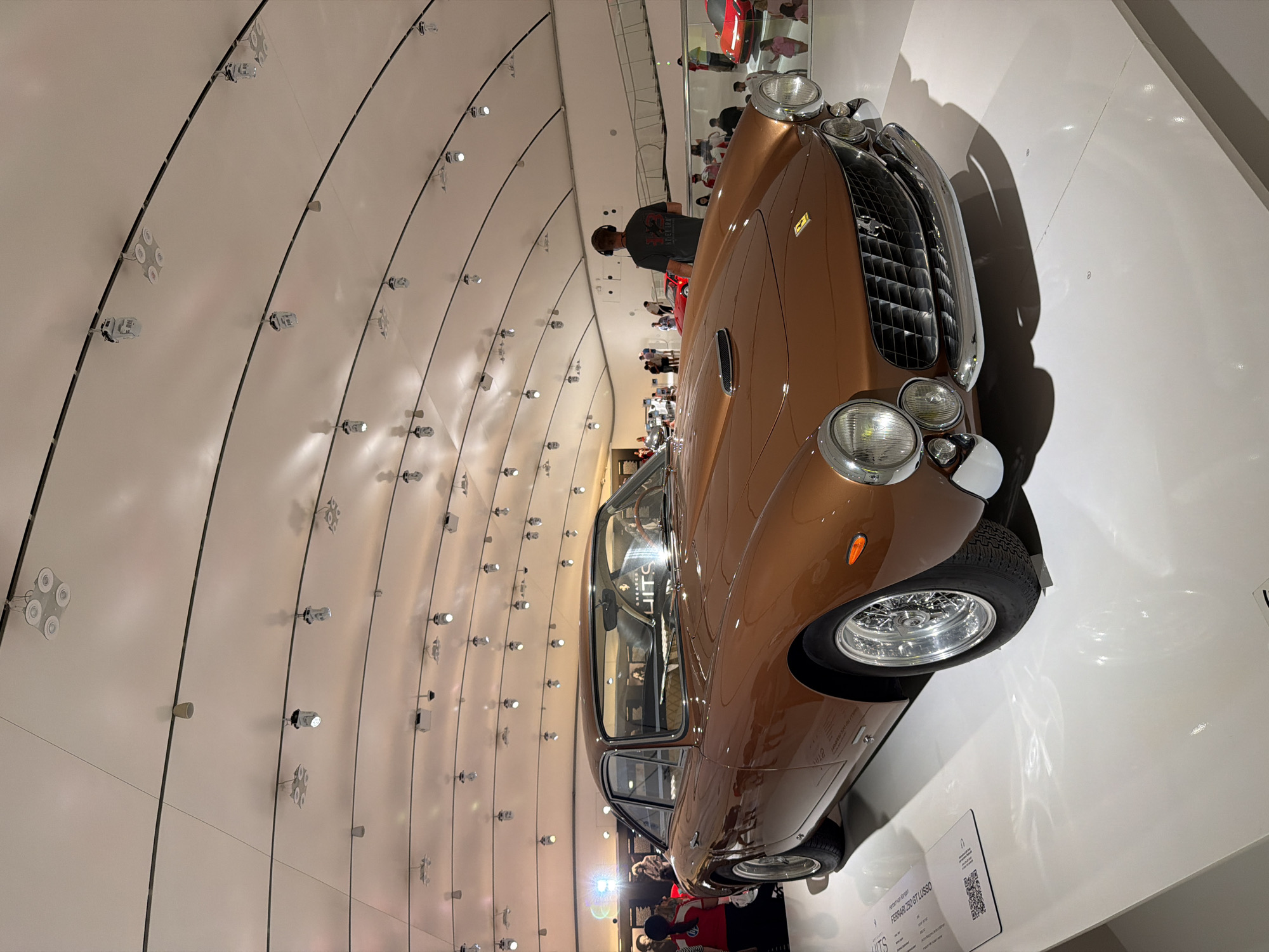 This image shows a vintage Ferrari 250 GT Lusso car on display in the Enzo Ferrari Museum in Modena, Italy. The car is a metallic bronze color and is positioned on a white, illuminated pedestal. The museum has a modern, curved ceiling with numerous track lights. Several people are visible in the background, some observing the car and others walking around the museum. The lighting is bright and designed to highlight the exhibits.