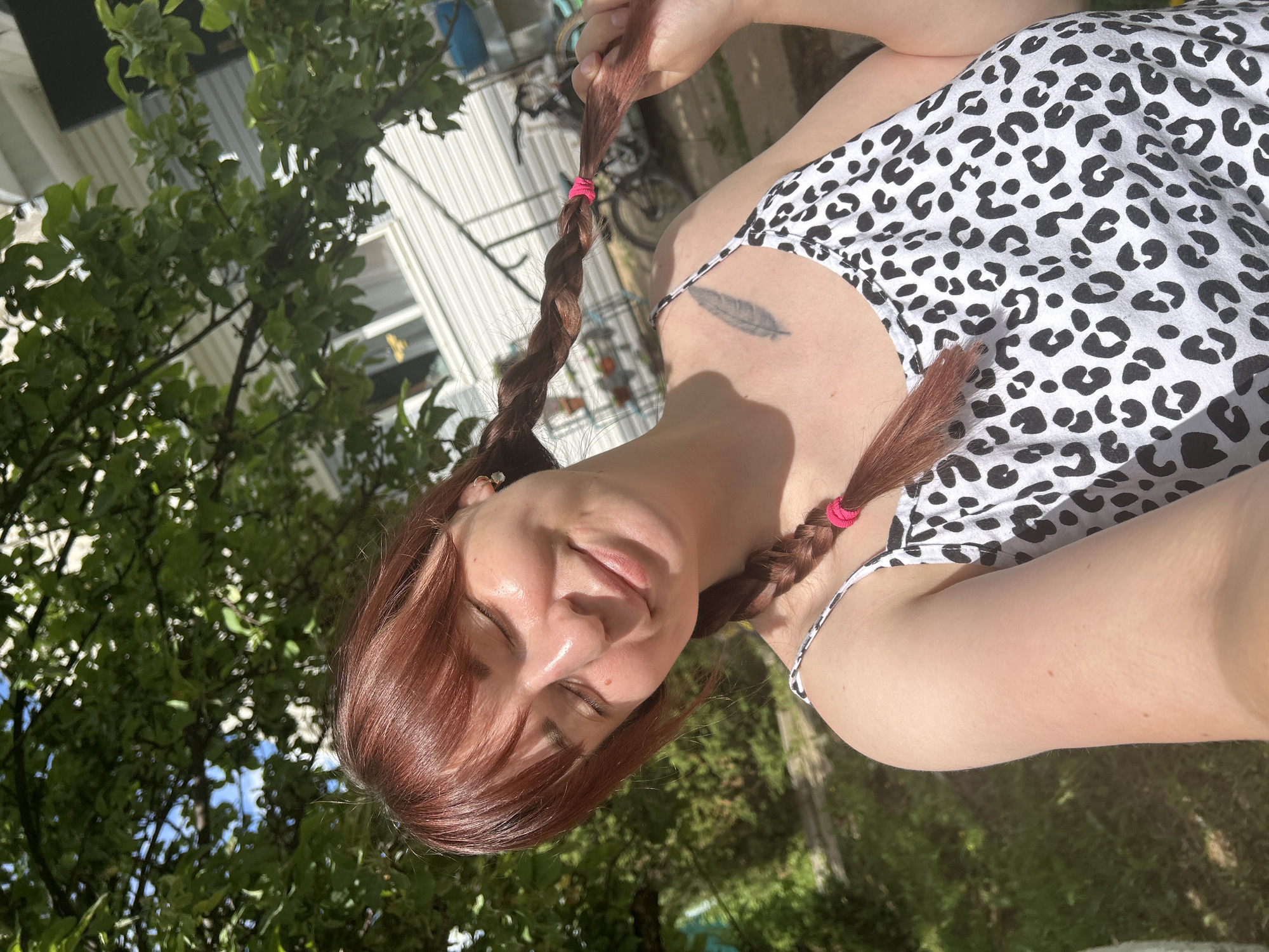 The image depicts a woman outdoors on a sunny day. She has reddish-brown hair styled in two braids secured with pink hair ties. She is wearing a white and black animal print top. A tattoo of a feather is visible on her upper chest. The background includes trees and a building. There is no indication that the location is Фоки, Russia.