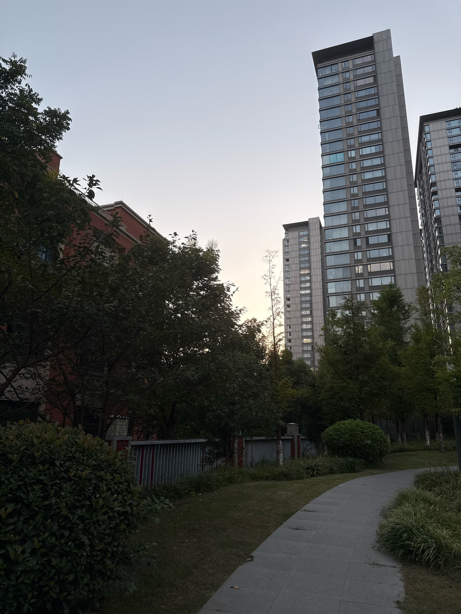 The image depicts a residential area in the city of Yiwu, China, as evidenced by the architecture and typical landscaping. In the foreground, a paved pathway curves through a grassy area with manicured shrubs and trees. A low fence with vertical slats separates the pathway from a green space with more trees and bushes. To the left, a red-brick building with white trim is visible, partially obscured by dense foliage. Dominating the background are several modern, tall apartment buildings with numerous windows. The sky is a pale, clear blue, suggesting either early morning or late evening. No people are present in the scene, and no actions are being depicted. The overall impression is one of a serene and well-maintained urban residential environment.