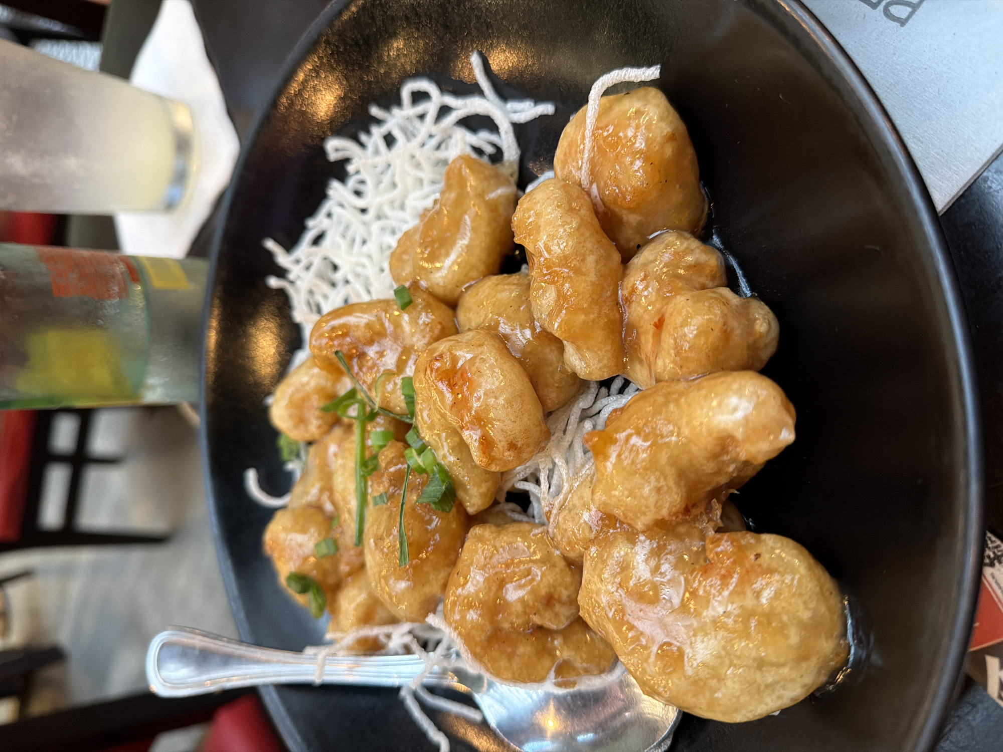 This image captures a close-up, top-down view of an appetizing dish served in a glossy black ceramic bowl, likely in a restaurant setting in Puebla, Mexico. The main subject is a generous portion of golden-brown, battered pieces of food, possibly shrimp or chicken, coated in a glistening, translucent light brown sauce that suggests a sweet and savory profile.
Beneath the main protein, a bed of finely shredded, crispy white strands, resembling fried rice vermicelli or thin noodles, provides a textural contrast. The dish is garnished with finely chopped green herbs, likely chives or green onions, adding a touch of freshness. A silver spoon is visible inside the bowl, resting among the food.
In the blurred background to the left, a tall, frosted glass containing a pale yellow liquid, possibly lemonade or a cocktail, stands beside a colorful bottle with red, green, and yellow bands. The dark table surface and indistinct shapes suggest an indoor dining environment. A small, partially visible piece of paper or card to the right of the bowl shows the upper part of the letter "P." No people are visible in the frame. The lighting is bright and direct, highlighting the inviting appearance of the meal.