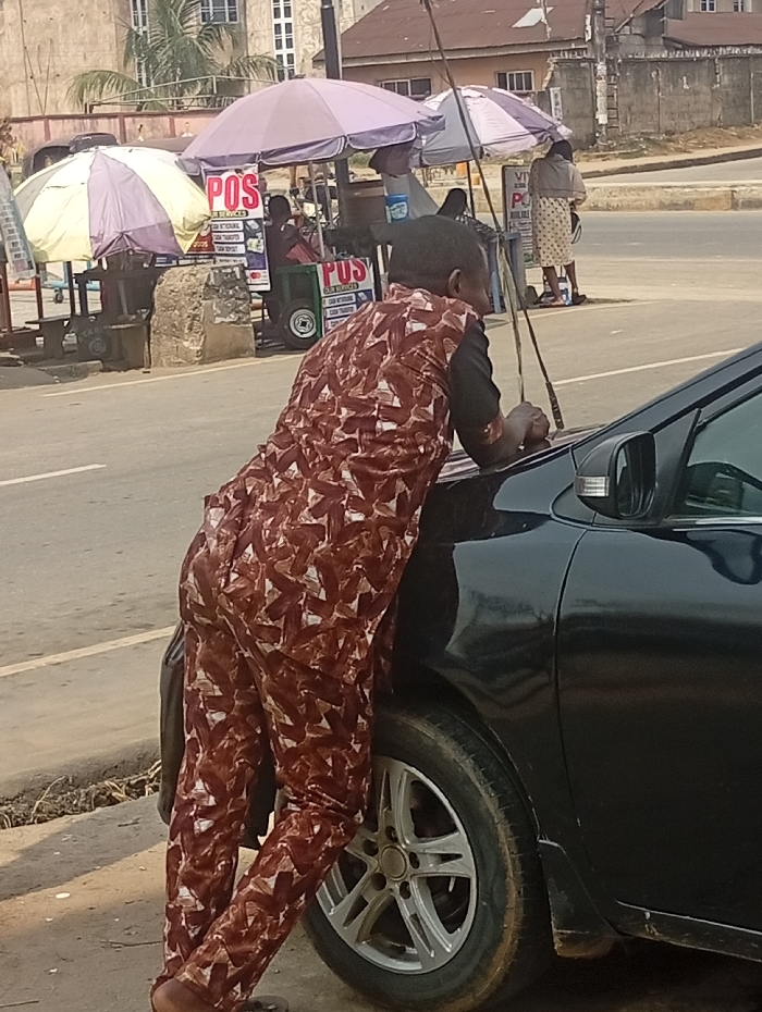 The scene depicts an urban street in Aba, Abia, Nigeria, under daylight conditions. In the immediate foreground, a person is positioned with their back mostly to the viewer, leaning forward against the front passenger side of a black sedan. The person wears a full-body garment patterned in reddish-brown and white, and holds a thin, dark stick-like object in their right hand, resting it on the car's hood. The black sedan, equipped with silver alloy wheels, is stationary on the paved roadside.
In the midground, directly behind the primary subject, a bustling roadside vending area is visible. Two large market umbrellas provide shade: one with yellow and white stripes to the left, and a light purple one to the right. Underneath these umbrellas, several individuals are present, and multiple signs displaying "POS" (Point of Sale) services are prominently featured. These signs advertise services such as "CASH DEPOSIT," "CASH WITHDRAWAL," and "CASH TRANSFER." A small green cart with wheels and another "POS" sign is visible under the purple umbrella. Further right, a person in a light-colored skirt or wrapper stands near a tall sign.
The background reveals multi-story concrete buildings and some palm foliage, indicating an urban setting. The overall environment appears dry and possibly dusty, consistent with typical street conditions.