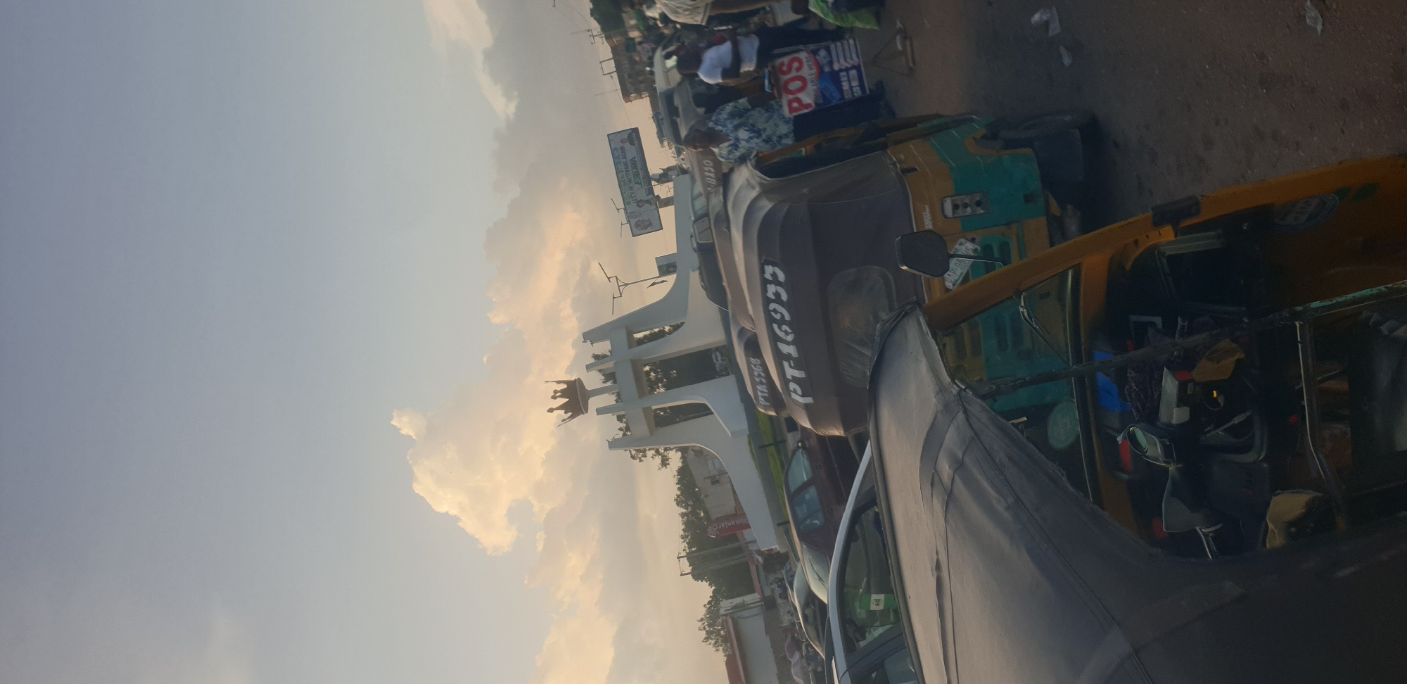 This media file shows a street scene in Barakin Lerawi, Nigeria, at what appears to be late afternoon or early evening. The sky is a pale blue with some fluffy, golden-tinged clouds, suggesting a warm and pleasant atmosphere. The scene is dominated by several auto-rickshaws, also known as Keke Napeps, which are common modes of transportation in Nigeria.
One auto-rickshaw in the foreground is primarily gray, with a black tarp covering part of it. Another in the mid-ground is a mix of dark brown and green, with yellow accents. A third auto-rickshaw, visible further back, is predominantly yellow. People are visible in and around the auto-rickshaws, suggesting they are either passengers or drivers. Some individuals are wearing casual clothing.
In the background, there is a large white monument with intricate designs, possibly a landmark or an architectural feature. A billboard or sign with text is also visible above the monument, and a smaller sign with "POS" is prominent on a blue background, indicating point-of-sale services. The road surface appears to be dirt or gravel. The overall impression is of a busy, active street with the hustle and bustle of daily life. The sunlight casts a soft glow on the scene, creating a warm ambiance. The orientation of the image is rotated, with the sky appearing on the left side of the frame.