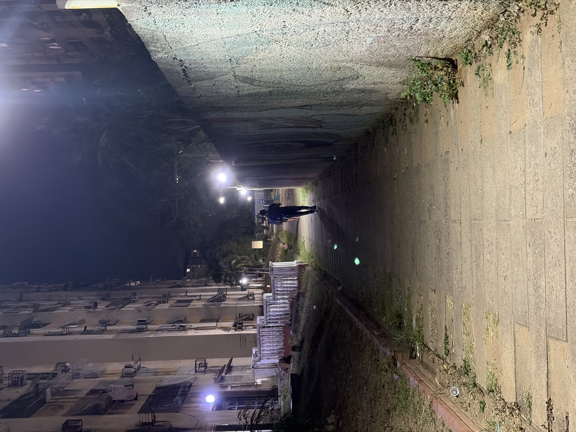 The image shows a person walking through an underpass in Ciudad Guayana, Venezuela. The underpass has concrete walls and a paved walkway. There are buildings to the left and vegetation along the right side. There are lights illuminating the walkway.