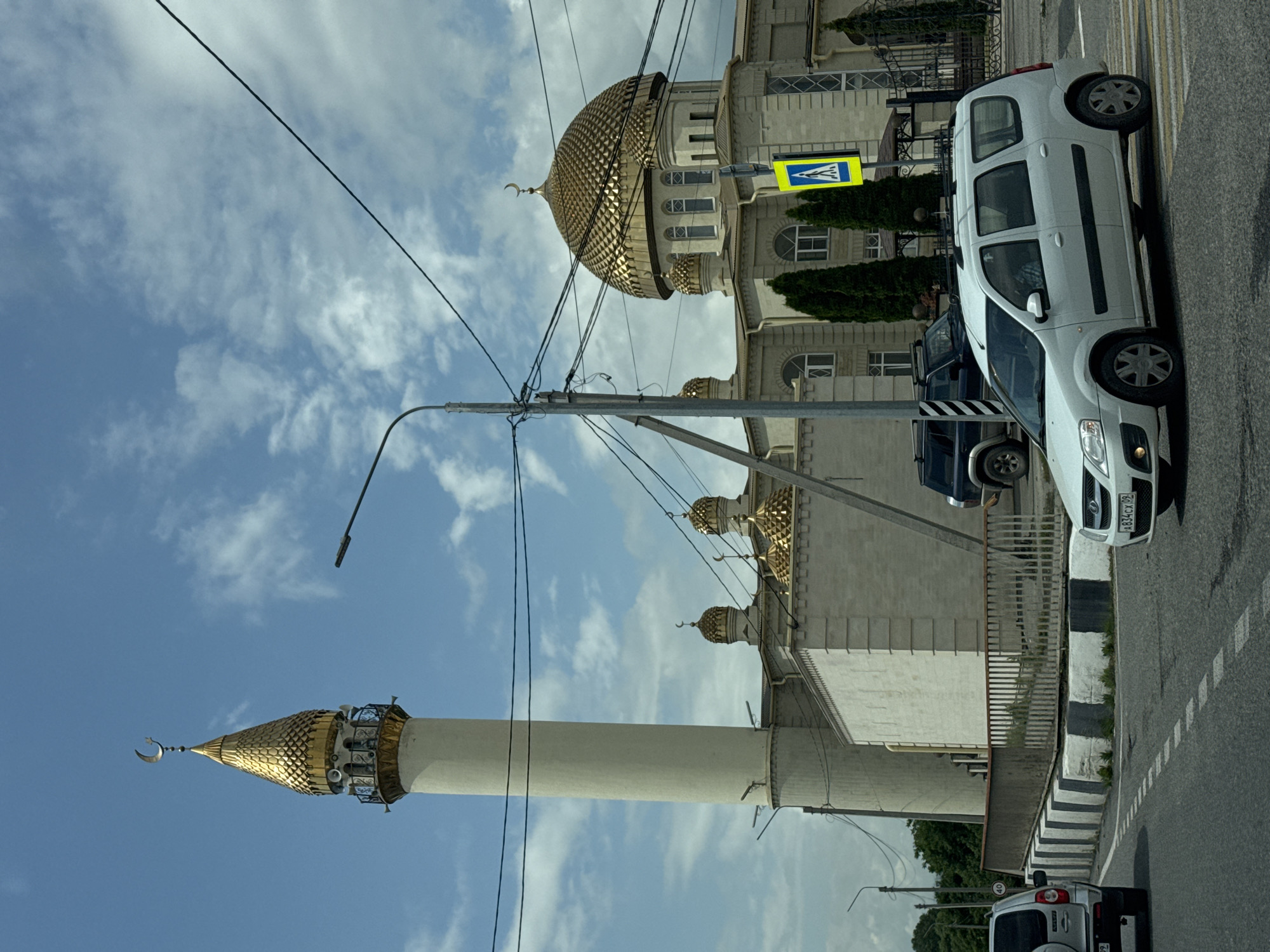 The image depicts a street scene in Ust-Dzheguta, Russia, prominently featuring a mosque with a tall, slender minaret topped with a golden, conical roof adorned with a crescent moon. The main mosque building has a large, golden dome also decorated with a crescent moon, and several smaller golden domes. The architecture is light-colored stone. In the foreground, a white station wagon is driving on a road with a pedestrian crossing. Other vehicles, including a dark SUV and another silver SUV, are visible. The environment includes trees, streetlights, power lines, and a clear blue sky with scattered clouds. A yellow pedestrian crossing sign is visible near the mosque entrance. The overall scene suggests a sunny day in a city.