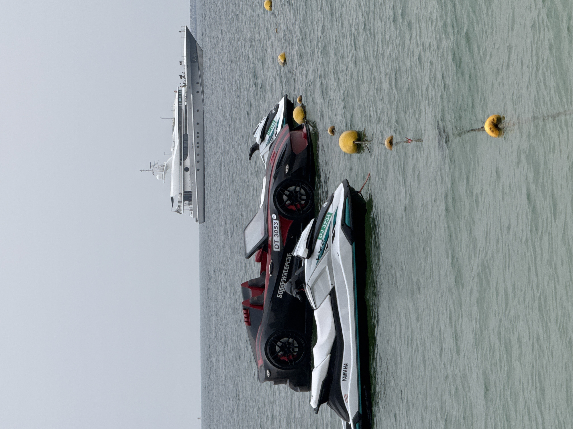 Two jet skis and a watercraft designed to resemble a sports car are floating in the sea. In the background, a large yacht is anchored. Several yellow buoys are anchored in a line, marking a boundary in the water. The sky is overcast and the sea is calm. The scene is likely in Dubai, United Arab Emirates, given the presence of a luxury yacht and high-end watercraft. No people are visible in the immediate vicinity of the watercraft, though there appear to be figures on the deck of the yacht. The watercraft are stationary and appear to be tied to moorings.