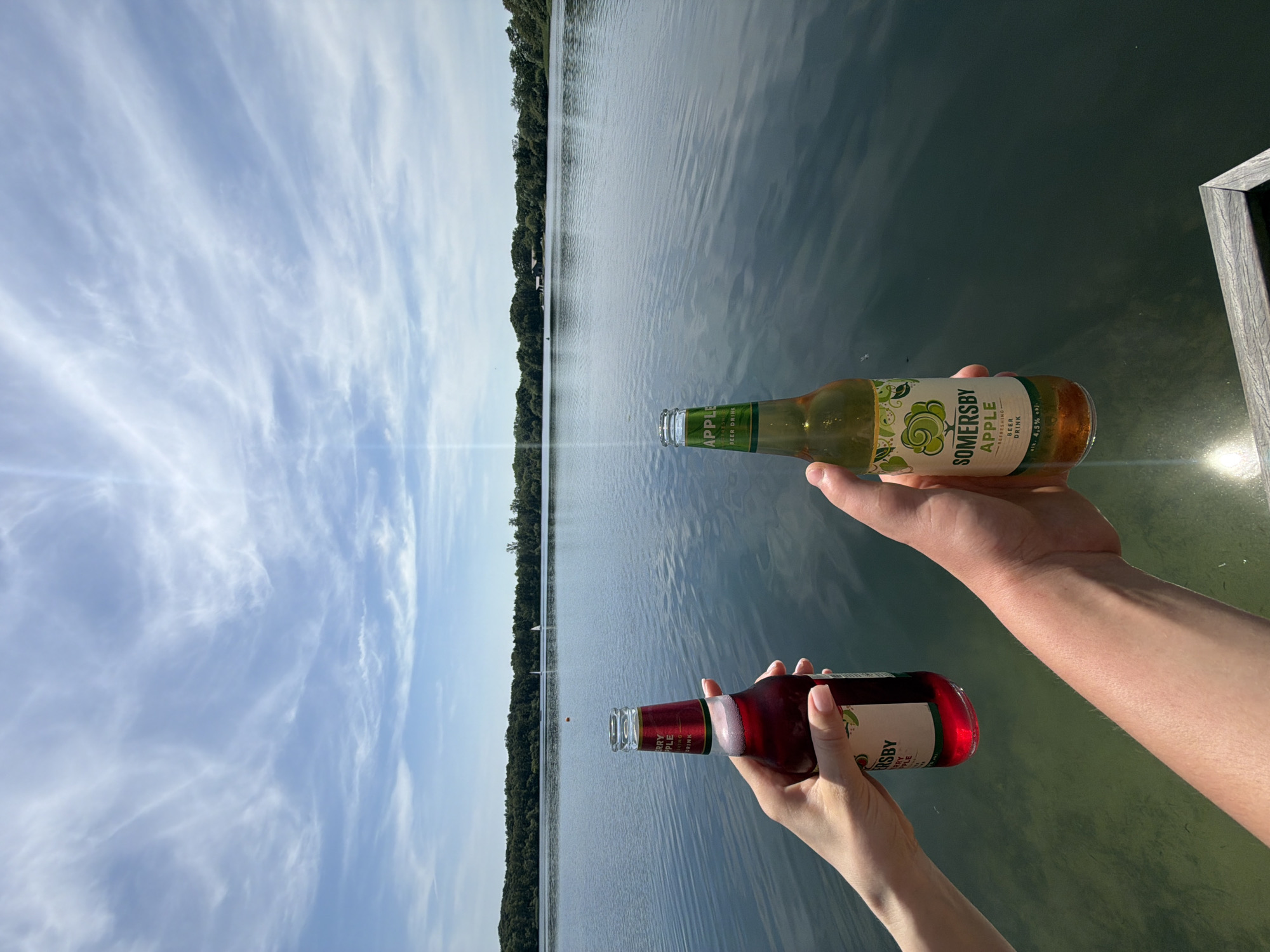 Two hands hold up bottles of Somersby Apple Beer Drink in front of a calm lake. The sky above is a clear blue with wispy white clouds. Trees line the far shore of the lake. The image was taken on a sunny day in Turnhout, Belgium.