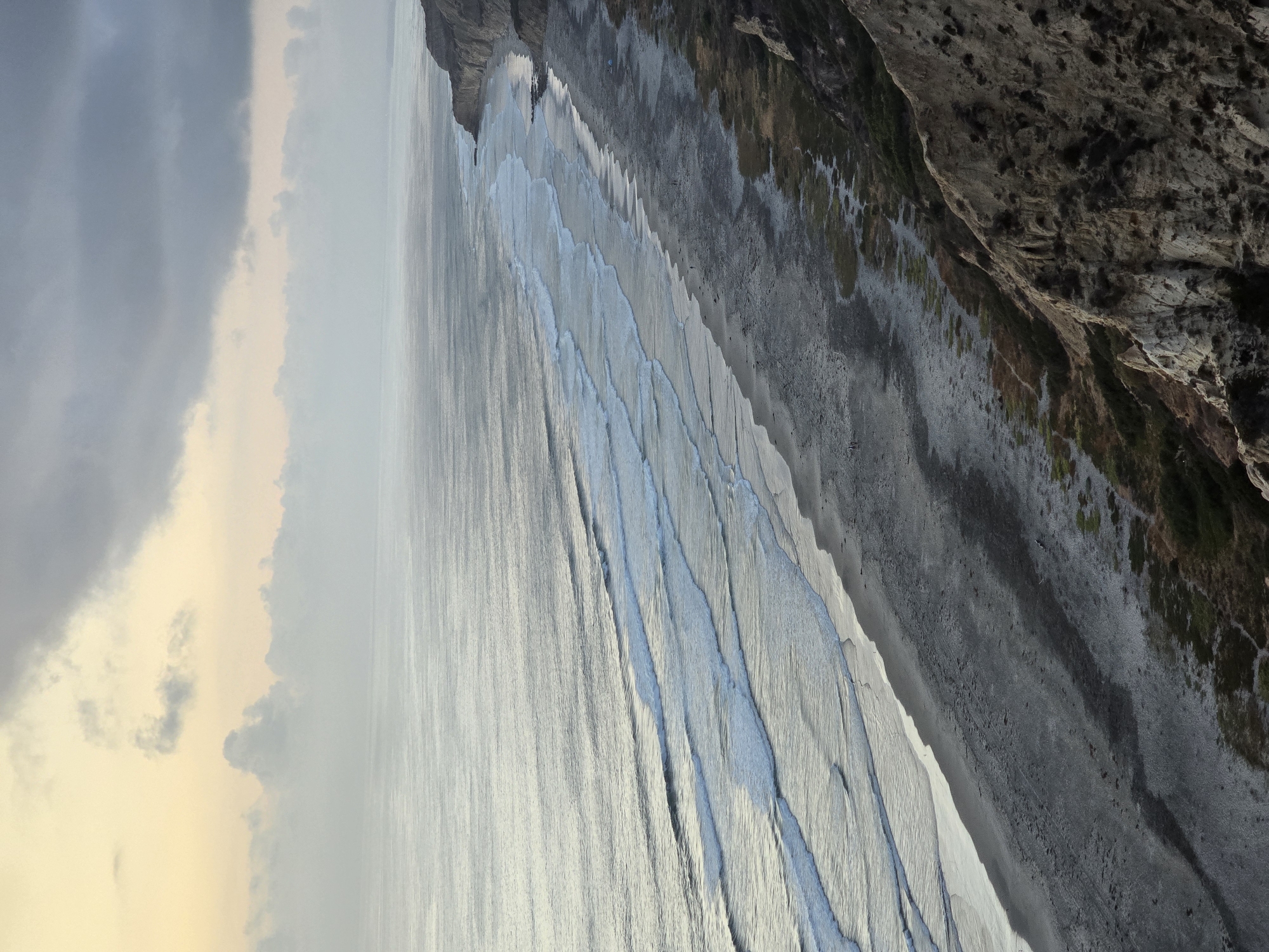 This is an aerial view of Torrey Pines State Beach in San Diego, California, on a cloudy day. The ocean stretches to the horizon, with gentle waves rolling onto the sandy shore. The beach is wide and appears mostly deserted, with a few scattered individuals walking along the waterline. To the right, the dramatic cliffs of Torrey Pines rise, covered in sparse vegetation. The sky above is overcast, with patches of pale yellow and gray clouds, suggesting either sunrise or sunset. The overall mood is serene and natural.