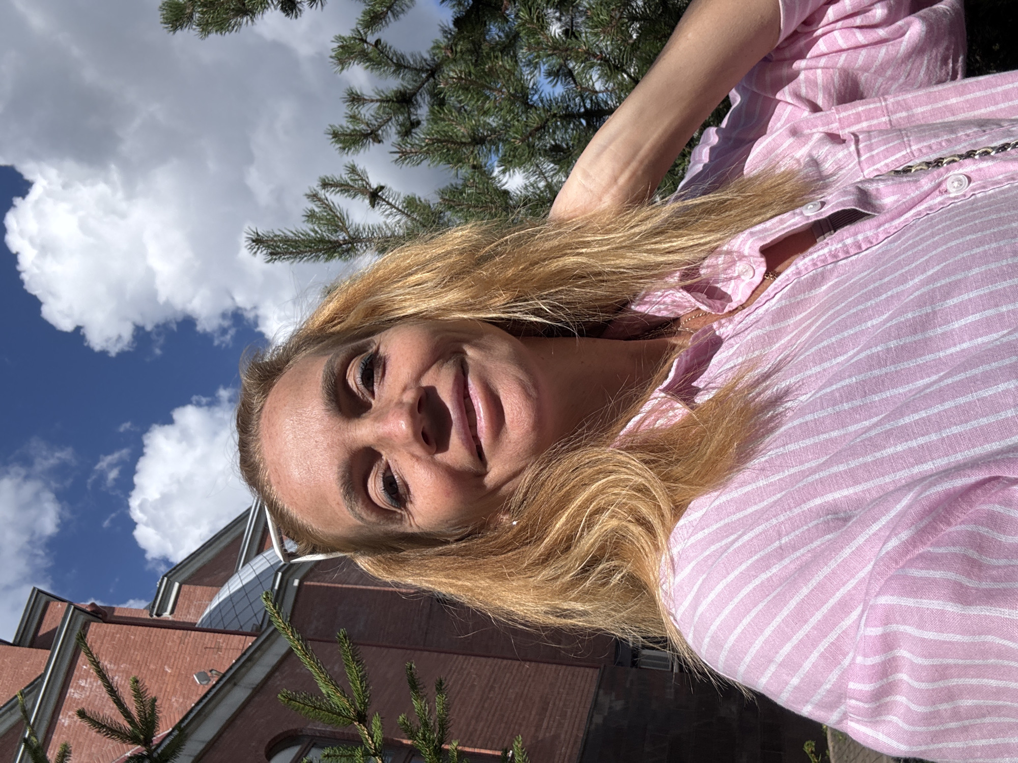 A woman with blonde hair is taking a selfie outdoors. She is wearing a pink and white striped shirt and a silver necklace. Her right arm is raised behind her head. In the background, there is a red brick building with a grey roof, green pine trees, and a blue sky with white clouds.