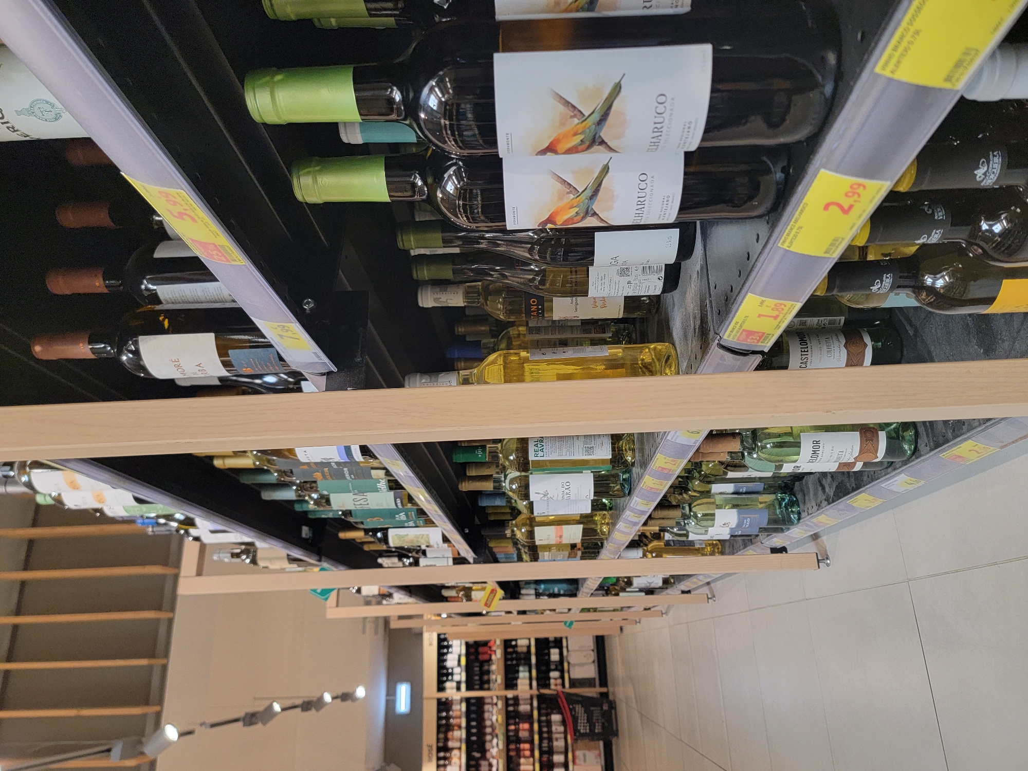The image shows a supermarket aisle filled with wine bottles on shelves. The bottles are mostly made of glass and vary in color, with some containing clear liquid and others dark liquid. Several bottles have labels with text and images, including some featuring birds. The shelves are made of metal and wood. Price tags are visible on some shelves, with prices like "1,89" and "2,99" marked in yellow. In the background, more shelves with bottles and what appears to be a shopping cart are visible. The lighting suggests an indoor commercial environment. The specific location in Faro, Portugal, is not identifiable from the image content.