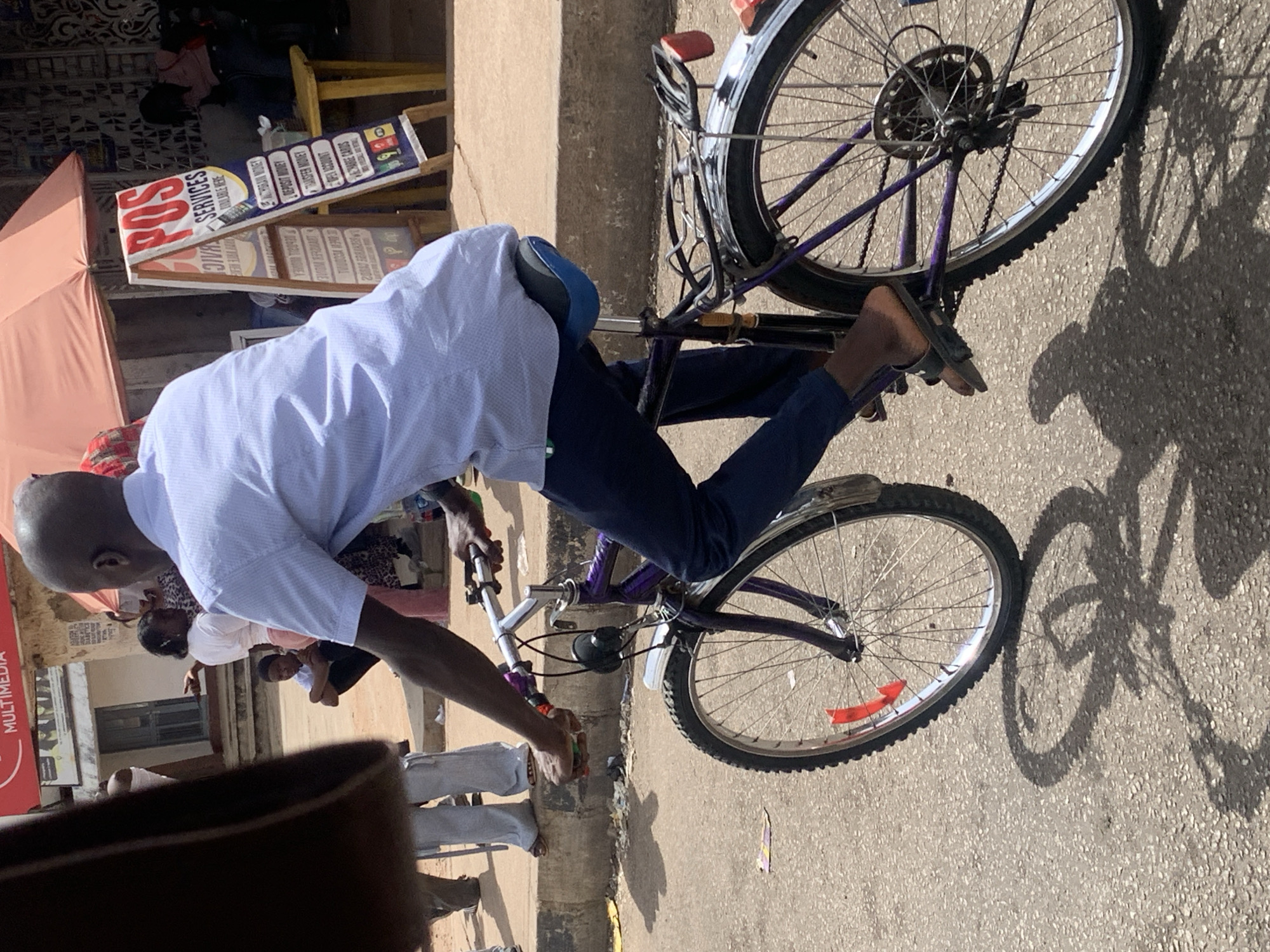 A man rides a bicycle in the city of Barakin Lerawi, Nigeria. The man is wearing a light blue shirt and dark pants. He is riding a purple bicycle on a paved road. The scene includes pedestrians, buildings, and a POS service sign. The environment is sunny with a visible shadow of the bicycle.