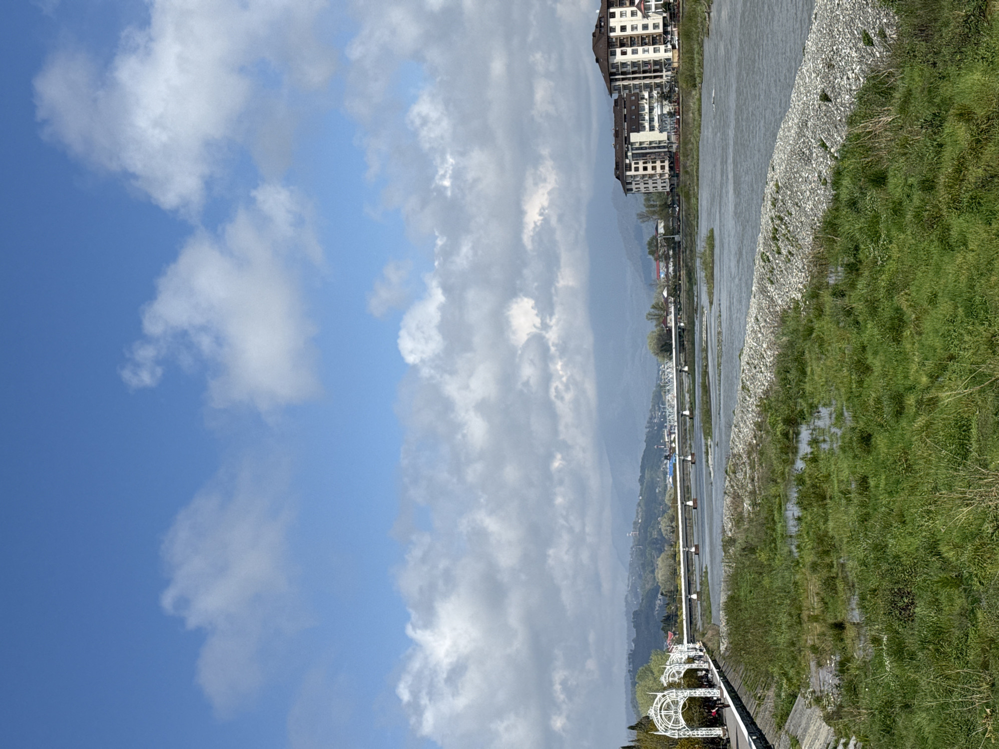 This panoramic landscape image captures a scenic view of a river and its surroundings in Sochi, Russia, under a partly cloudy sky. The upper portion of the frame features a clear blue sky interspersed with numerous fluffy white and grey clouds. In the background, hazy, light grey mountains rise, indicating the region's mountainous terrain.
The scene is dominated by a wide river that flows from the background towards the foreground. Its right bank is characterized by a stretch of light-colored stones or pebbles, transitioning into a lush green grassy area that extends to the far right of the frame. Some parts of the grassy bank appear wet or marshy near the water's edge.
On the left side of the river, several modern multi-story buildings with balconies and varied facades are visible, suggesting a developed urban or resort area. These structures line the riverbank. In the foreground on the left, a paved promenade or pathway runs parallel to the river, featuring elegant white decorative architectural elements, including ornate archways or gazebos, one of which has a prominent circular design. A long, low-lying structure, possibly a pedestrian bridge or a series of piers with white railings, extends along the left side of the river into the distance.
The lighting indicates it is daytime, likely mid-morning or afternoon, with bright conditions despite the clouds. No people or visible text are discernible. The overall atmosphere is tranquil, highlighting the blend of natural beauty and urban development characteristic of a resort city like Sochi.