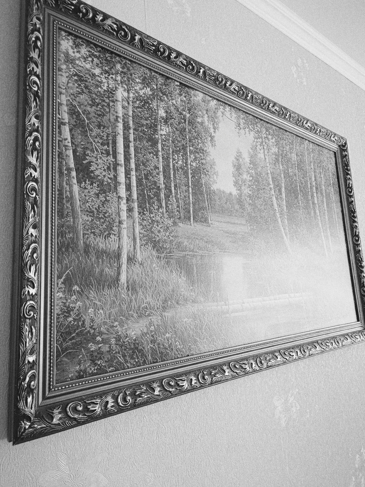 A black and white photograph depicts a framed picture hanging on a wall. The picture within the frame appears to be a landscape scene featuring birch trees, grass, and possibly a body of water, such as a stream or pond. The frame is ornate and decorated with scrolling patterns. The wall is covered in textured wallpaper with a subtle floral pattern. Based on the style of the wallpaper and frame, the image could be from a residential interior. No people or actions are apparent in the scene. The photo does not provide enough information to confirm whether it is located in Tobolsk, Russia.