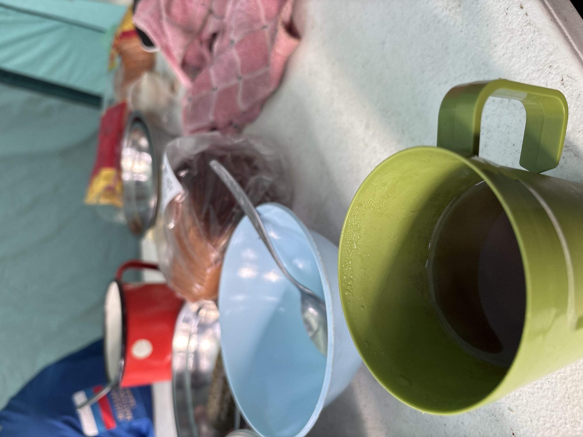 A top-down, slightly angled view captures an assortment of casual kitchenware and personal items arranged on a light-colored, possibly white or off-white, surface. In the immediate foreground to the right, a bright lime green plastic mug with a handle holds a dark liquid, likely a hot beverage like coffee or tea, with condensation droplets visible on its inner wall.
To the left of the green mug, a light blue plastic bowl contains a silver spoon. Behind these, items are stored in clear plastic bags, possibly containing food such as bread. Further to the left, a red metal mug with a white circular marking is partially visible, alongside several stacked metallic bowls or cups.
In the upper background, a pink patterned cloth or towel is laid out. The left side of the image features a distinct light blue or teal fabric, strongly suggesting the interior wall of a tent or similar outdoor shelter. Towards the bottom left, a blue fabric item, possibly clothing or a bag, displays a label with indistinct text and red, white, and blue stripe details.
The surface itself appears slightly worn or speckled, common in outdoor settings. The overall scene suggests a break for a meal or refreshments during a camping trip or an outdoor activity in Lesnoye, Russia. No individuals are present, and the lighting does not provide clear indications of the time of day or external weather.