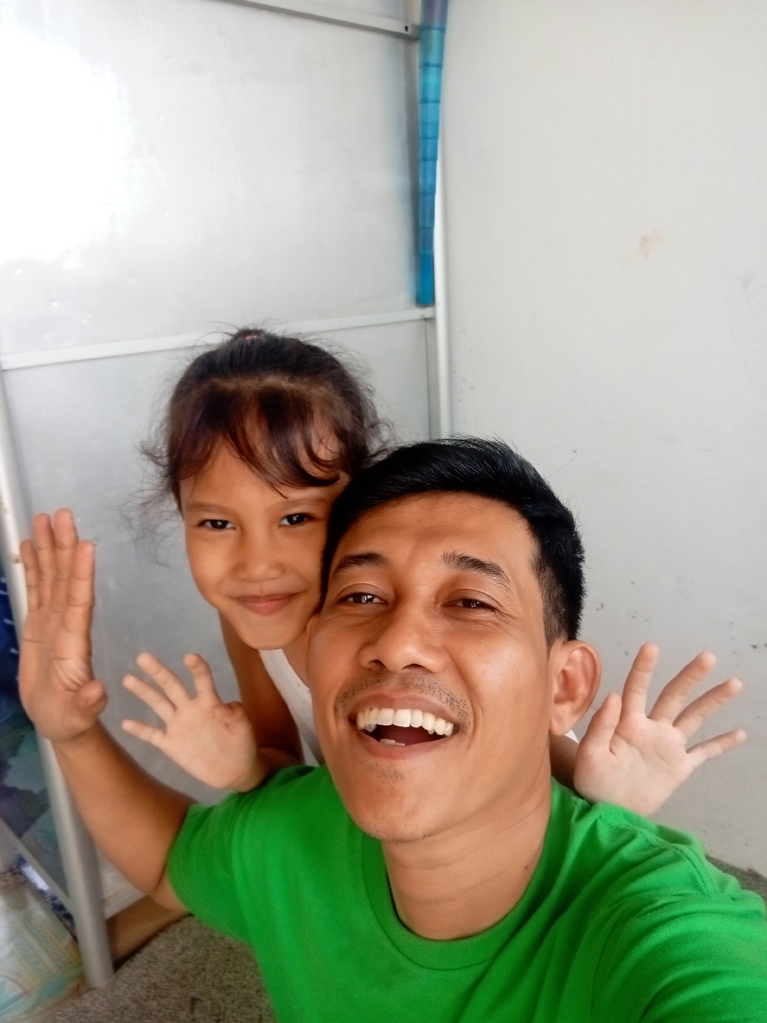 In Batam, Indonesia, a selfie is taken by a man wearing a green t-shirt, who is smiling broadly and looking directly at the camera. Behind him, a young girl with her hair pulled back in a bun is peeking over his shoulder, also smiling. Both are raising their hands in a playful gesture, as if waving. The background consists of a plain, light-colored wall and a metal frame structure, suggesting an indoor setting. The lighting is bright, with some glare on the wall, indicating it might be daytime.