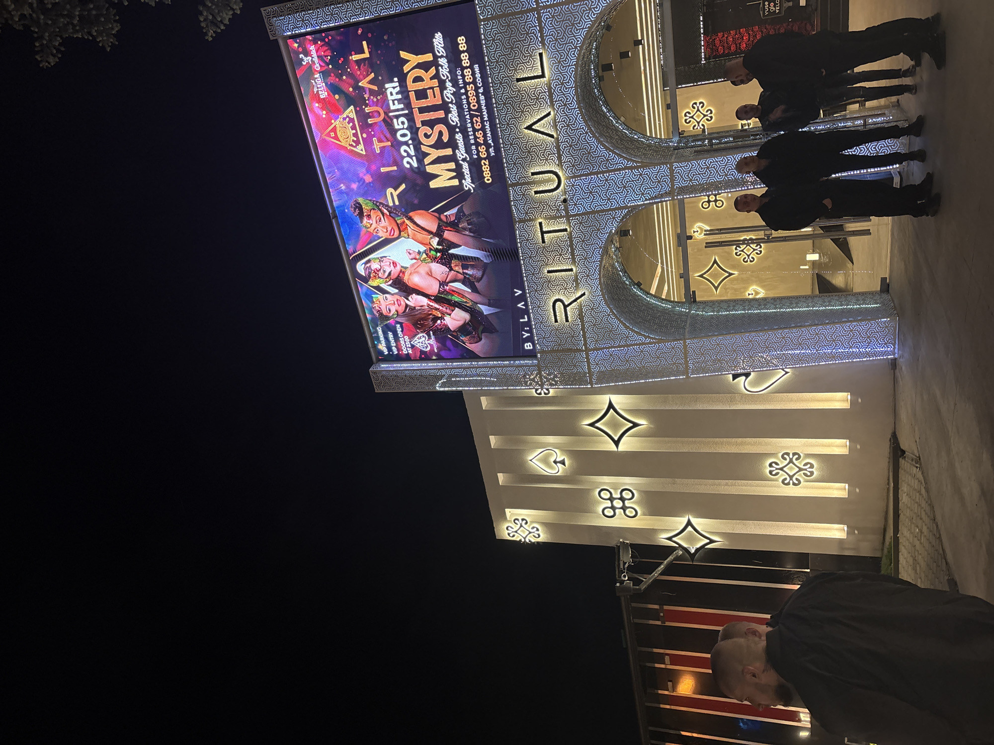 Here is a description of the image:
The image depicts the exterior of a building at night, likely a club or entertainment venue named "RITUAL," located at УЛ. „АТАНАС МАНЧЕВ" 6, СОФИЯ (Sofia). The building's façade is illuminated and features ornate decorations. A large digital billboard displays promotional content for an event on 22.05 (May 22nd) titled "MYSTERY," featuring the text "Special Guests Best Pop-Folk Hits" and contact information for reservations. Decorative elements on the building include lit-up card suit symbols (spade, diamond, club, heart). Several people, likely patrons or staff, are standing in front of the building's entrance. One person, with a shaved head, is closer to the foreground. The overall atmosphere suggests an upscale nightlife establishment.