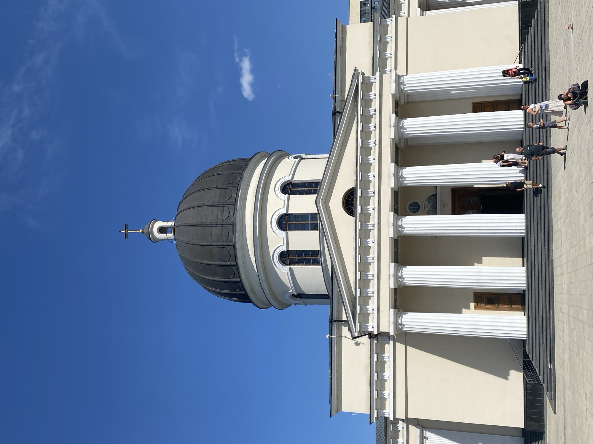 The image shows the Metropolitan Cathedral of the Nativity in Chișinău, Moldova. It is a neoclassical cathedral with a large dome topped by a cross. The facade is adorned with white columns and decorative molding. Several people are visible in front of the cathedral, some ascending the stairs, while others are walking or standing on the paved plaza. The sky is a clear blue with a few wispy clouds. The lighting suggests it is daytime, likely sunny.