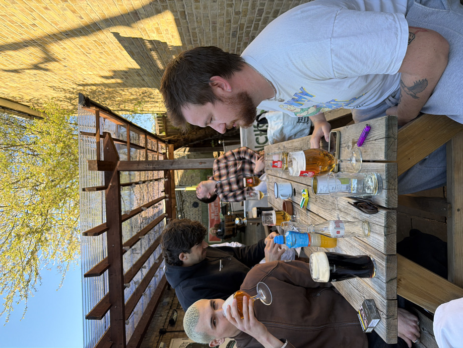 A group of four young adult males is depicted in an outdoor pub garden setting, likely "The Albert" in London, United Kingdom. The scene is illuminated by strong, low-angle sunlight, casting distinct shadows of a pergola structure onto a yellow-brick building in the background.
The individuals are seated around a rustic wooden picnic table laden with beverages and personal items. From left to right:
1. A male with short, bleached blonde hair, wearing a brown jacket, is shown bringing a stemmed glass of amber-colored beer to his mouth.
2. A male with dark, curly hair, dressed in a black hoodie, leans forward over the table, his hands positioned near a blue plastic bottle.
3. A male wearing a plaid shirt is seated with his head tilted sharply upwards, looking towards the sky.
4. A male with brown hair and a beard, wearing a white t-shirt and grey sweatpants, is seated on the right side of the table, facing left. A tattoo of a bird is visible on his right forearm.
On the wooden table, the following items are present: a pack of Marlboro cigarettes, a pint glass containing a dark stout beer, an empty clear glass bottle, a small glass of orange liquid, a blue plastic water bottle, a pair of black sunglasses, multiple beer glasses (some full, some partially consumed, containing amber-colored lager), a white enamel mug, several metal cans, a mobile phone, and a purple cigarette lighter. Above the table, a pergola with a corrugated clear plastic roof is visible. A tree with bright green foliage and a clear blue sky form the upper background.
